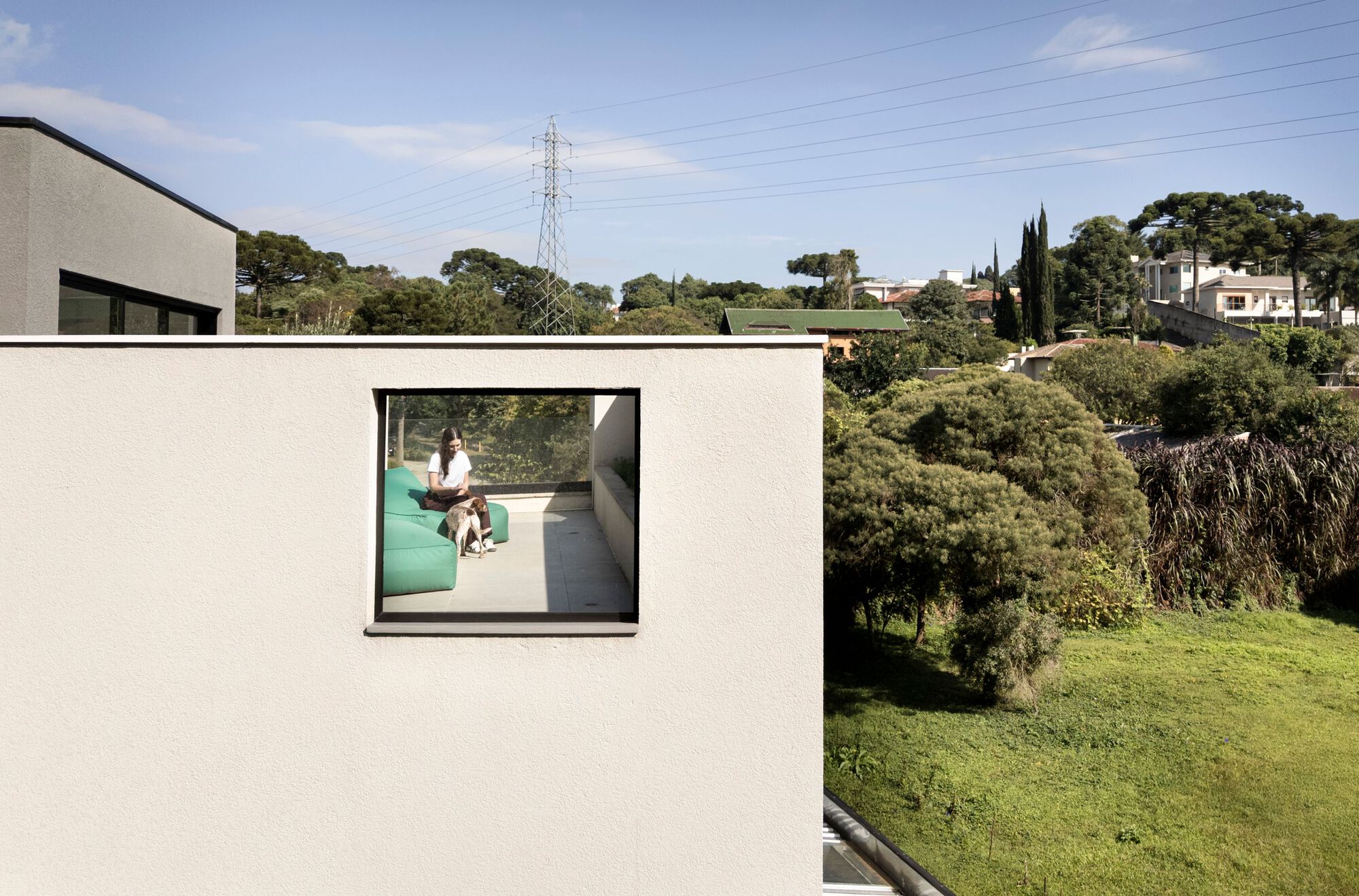 Square window in a white rendered wall framing a woman and dog on a green sofa, with trees and pylon beyond