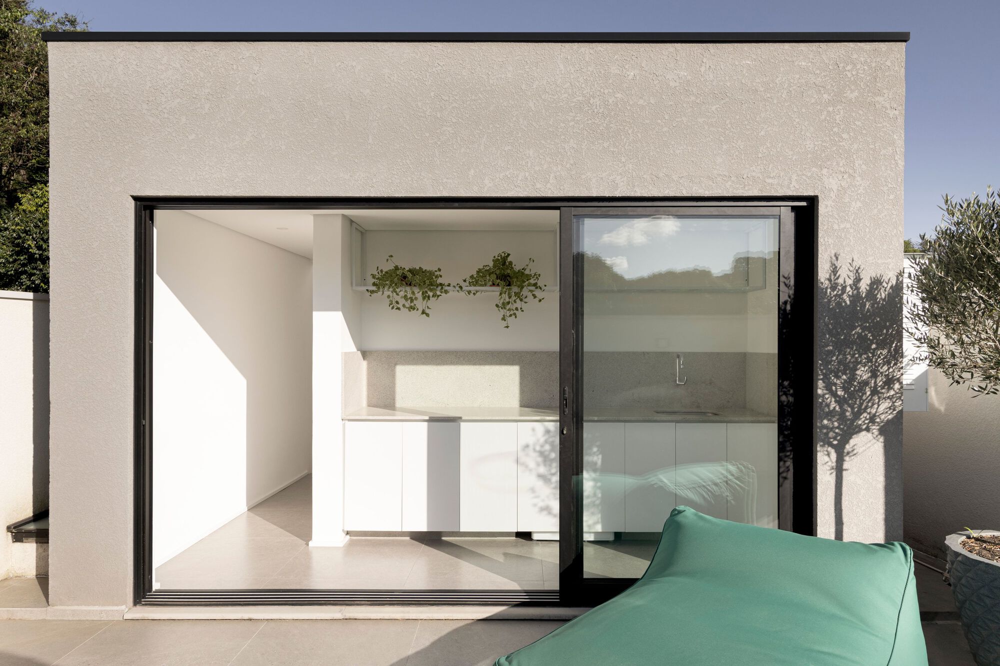 Sliding glass door opening onto a white kitchenette with hanging plants, seen past a green bean bag in afternoon sun
