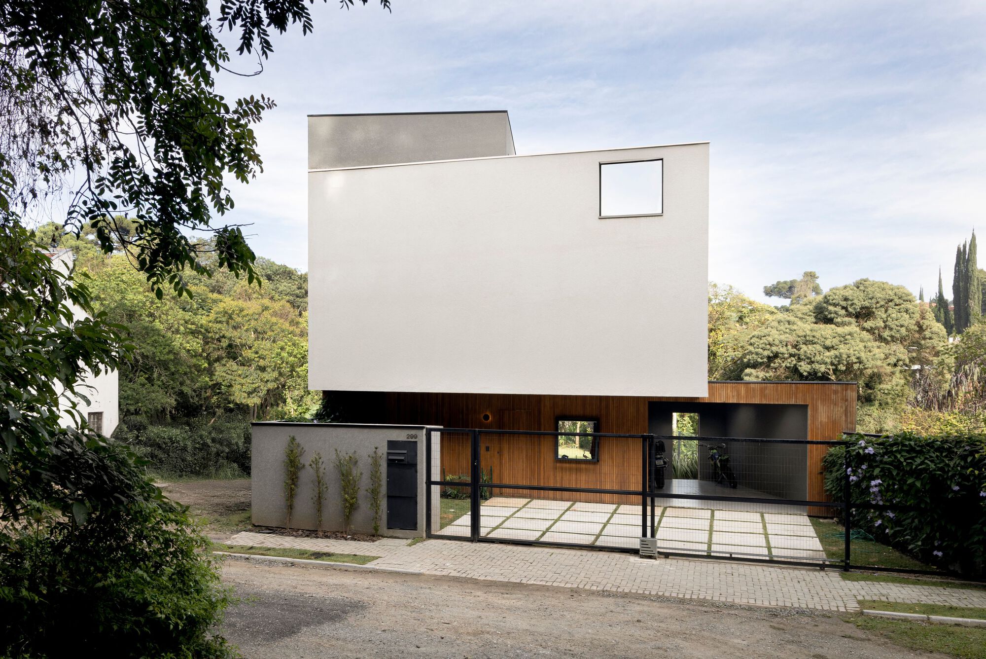 Street view of the white cantilevered upper volume above a timber-clad base and black metal gate, framed by trees