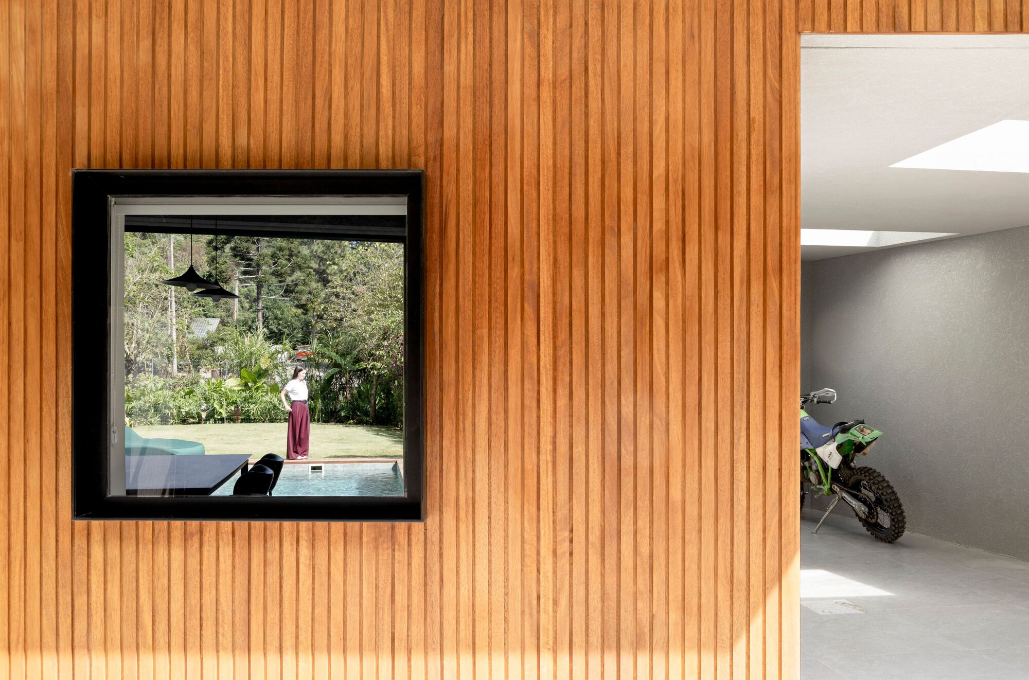 Vertical timber slat wall with a square black-framed window reflecting a woman by the pool, and a green dirt bike parked in the garage beside it