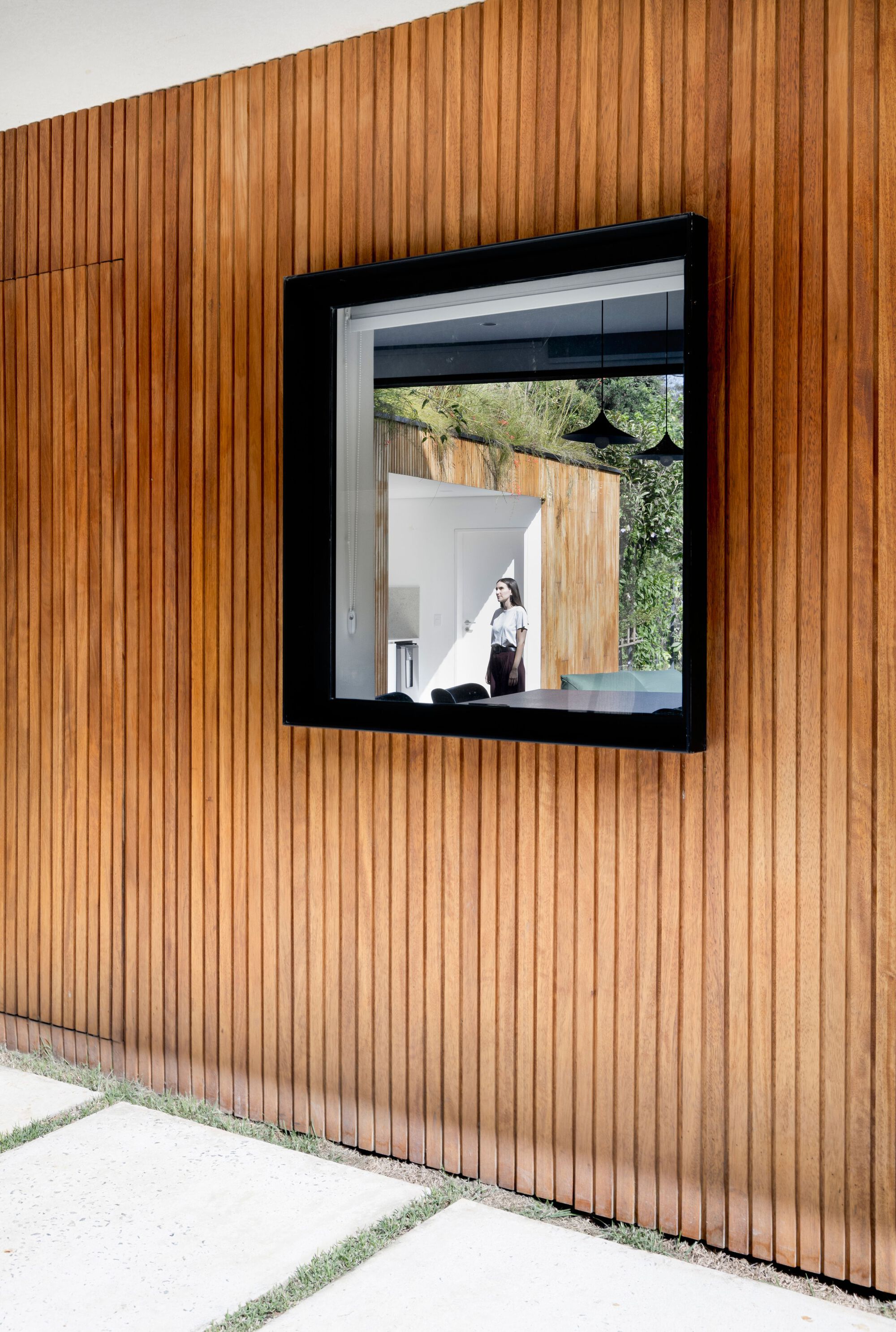 Square black-framed window set in vertical timber cladding, with a person visible inside and concrete pavers set in grass below