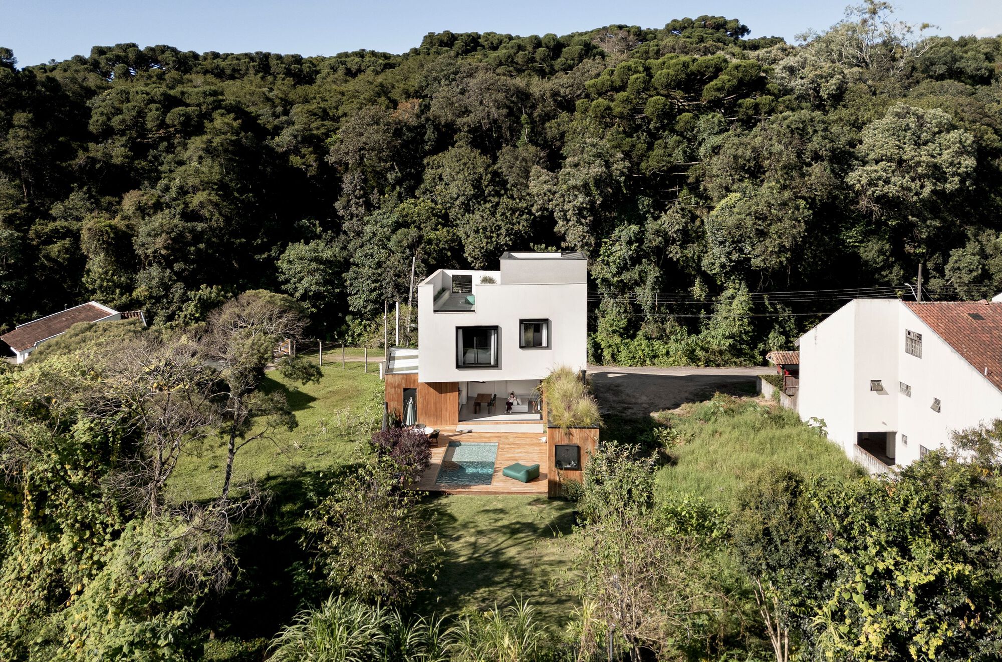 Aerial view of the white stacked house with a timber deck and small pool, set against a dense forested hillside