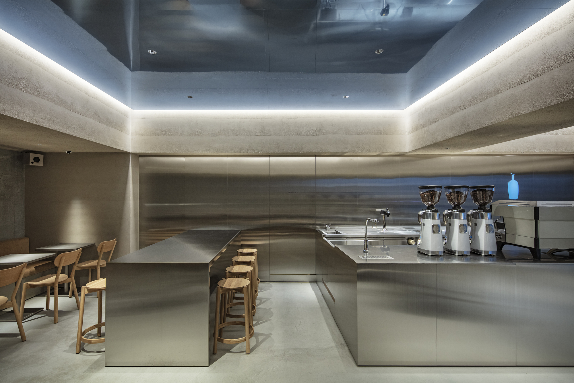 Stainless steel counter and wooden stools beneath a mirrored ceiling recess with cove lighting, three grinders lined up on the bar
