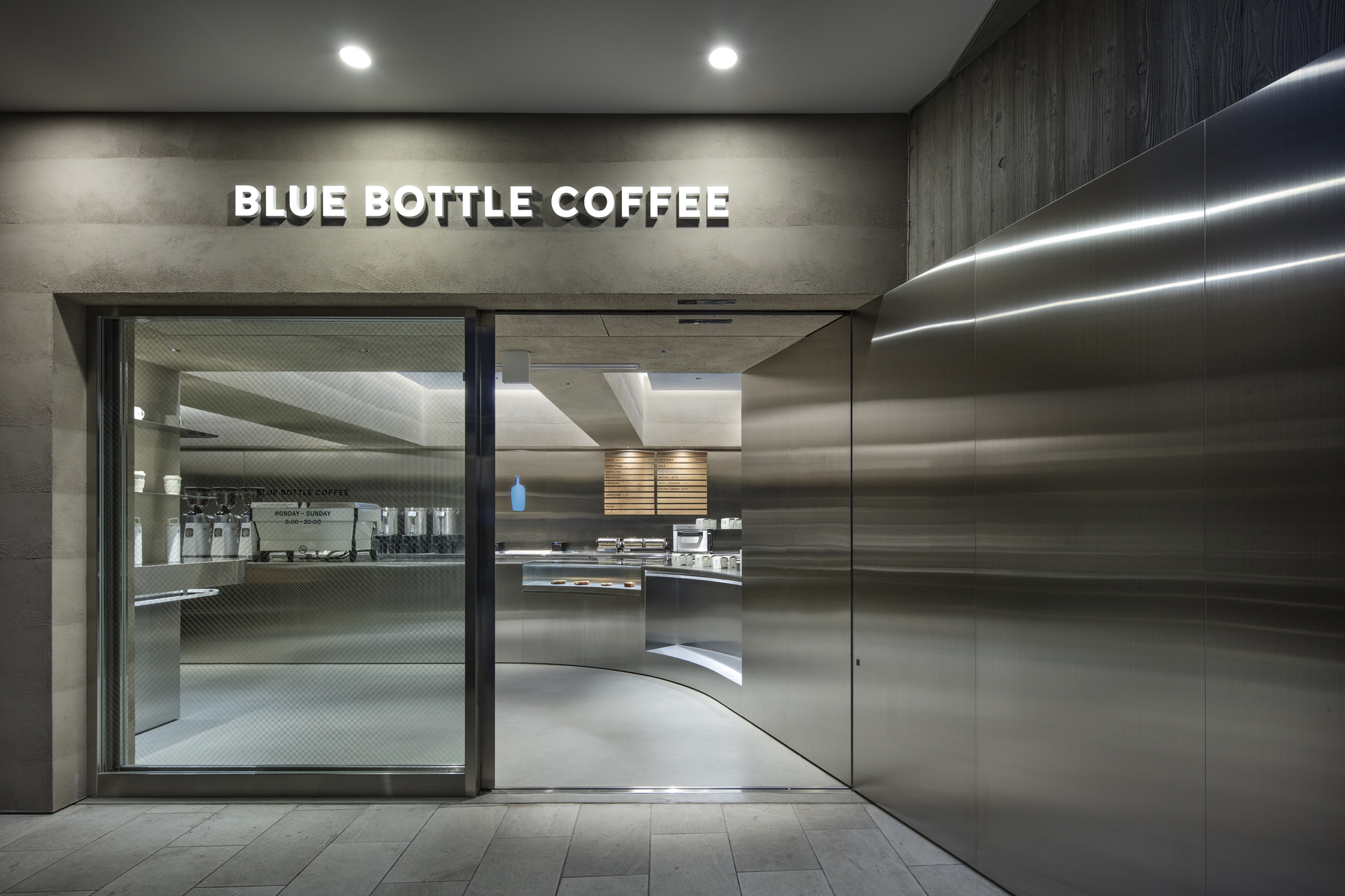 Shopfront entrance with glass door and curved stainless steel wall, lit strips reflecting along the metal at dusk