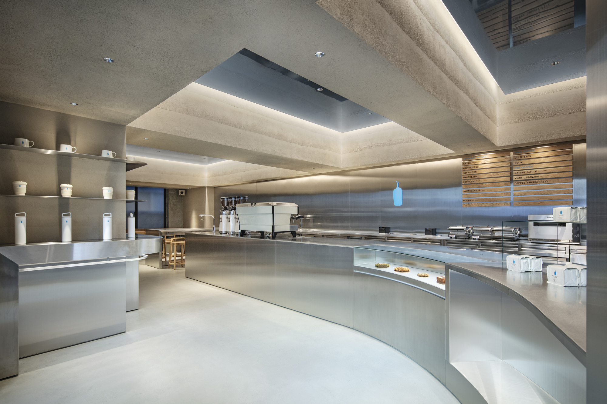 Curved stainless steel counter with pastry display case, wall shelves of cups and a stepped concrete ceiling glowing with cove light
