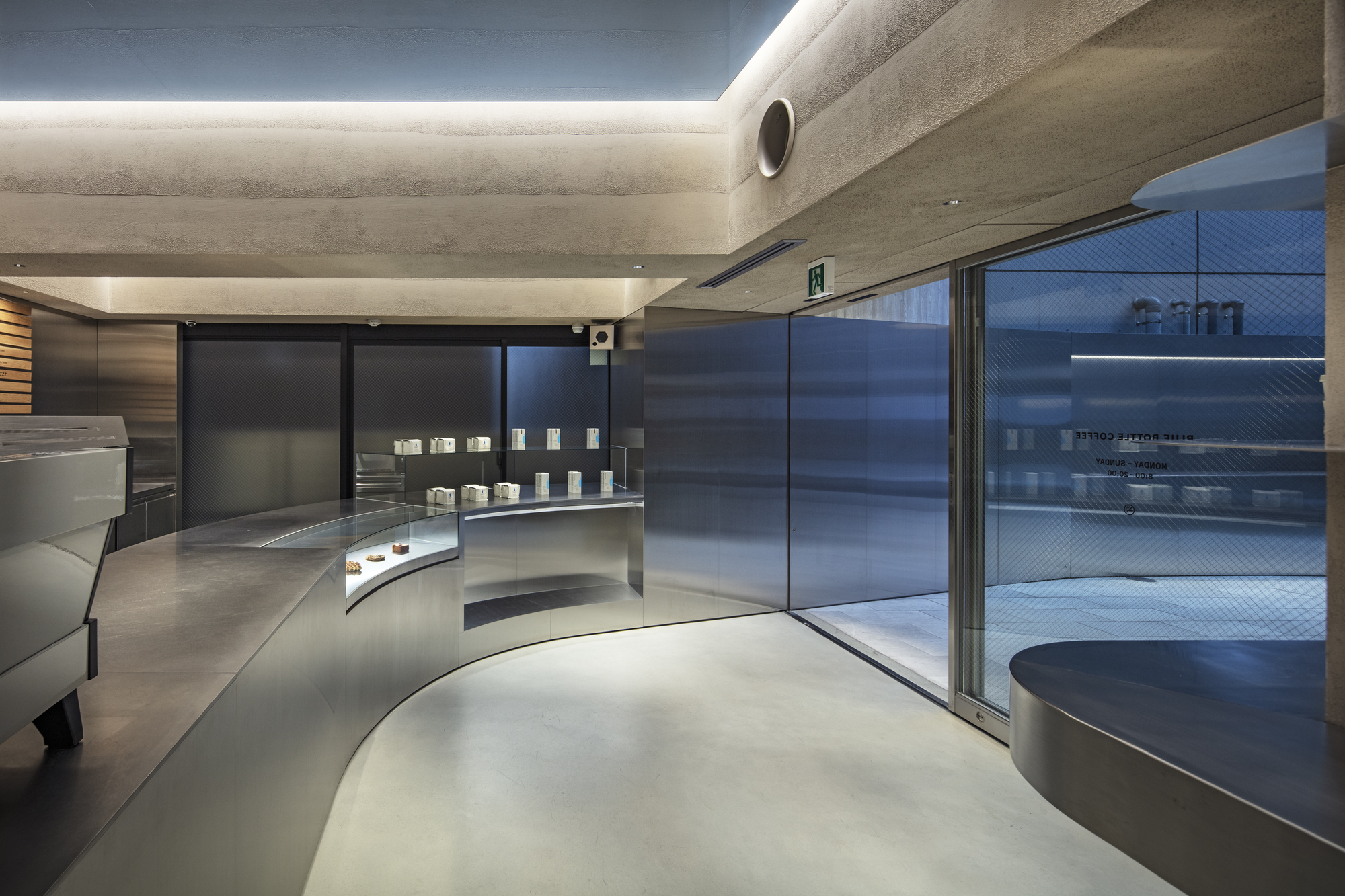 Sweeping steel counter curving toward the glass entrance, with a recessed pastry case and a rough concrete ceiling edge lit from above
