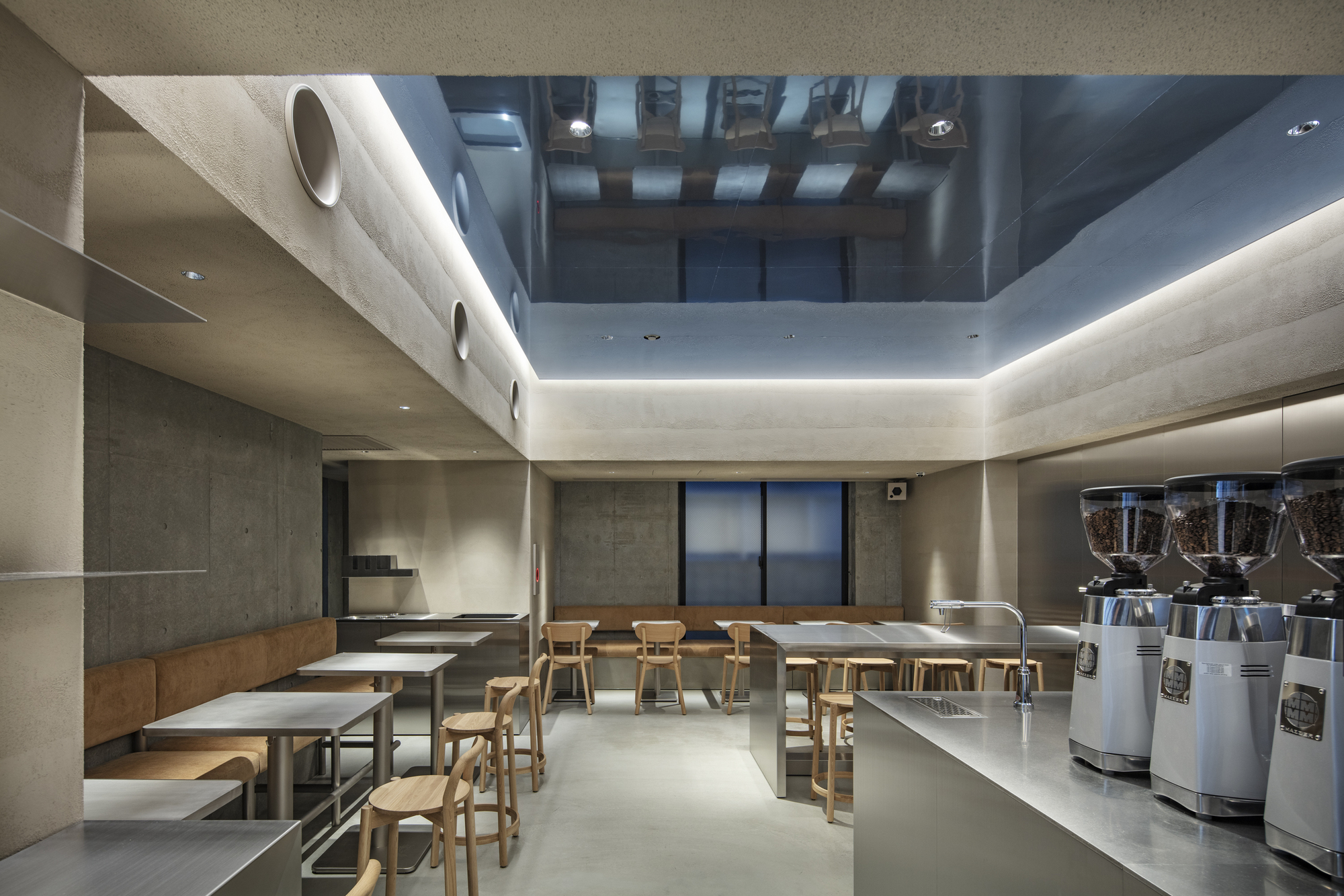 Seating area with wood banquette and tables below a mirrored ceiling well, grinders on the steel counter in the foreground