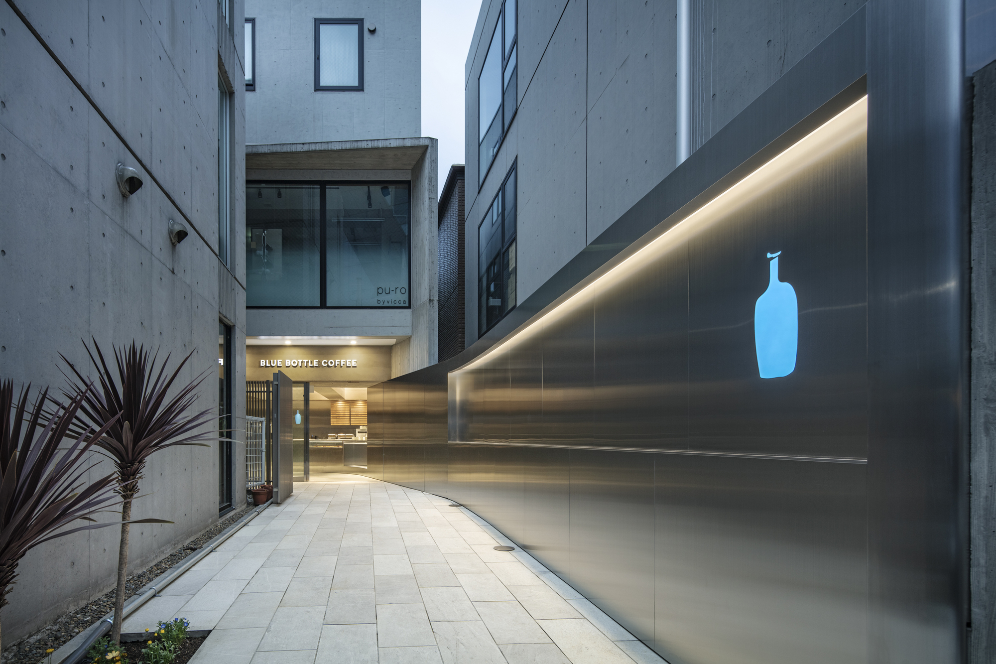 Curving stainless steel wall with glowing blue bottle logo along a paved alley, red-leaved plants beside the concrete facade at dusk