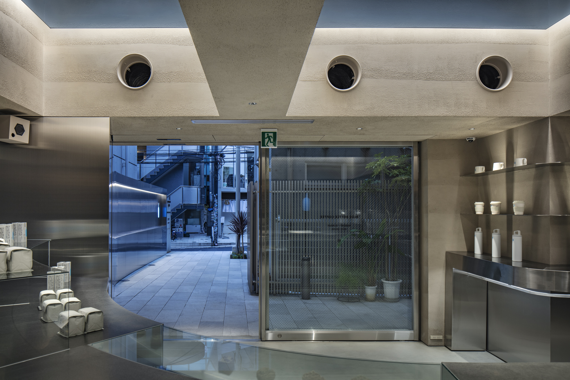Glass entry door opening to a paved courtyard with potted plants, seen past stainless steel counters and round vents in the rammed earth wall at dusk