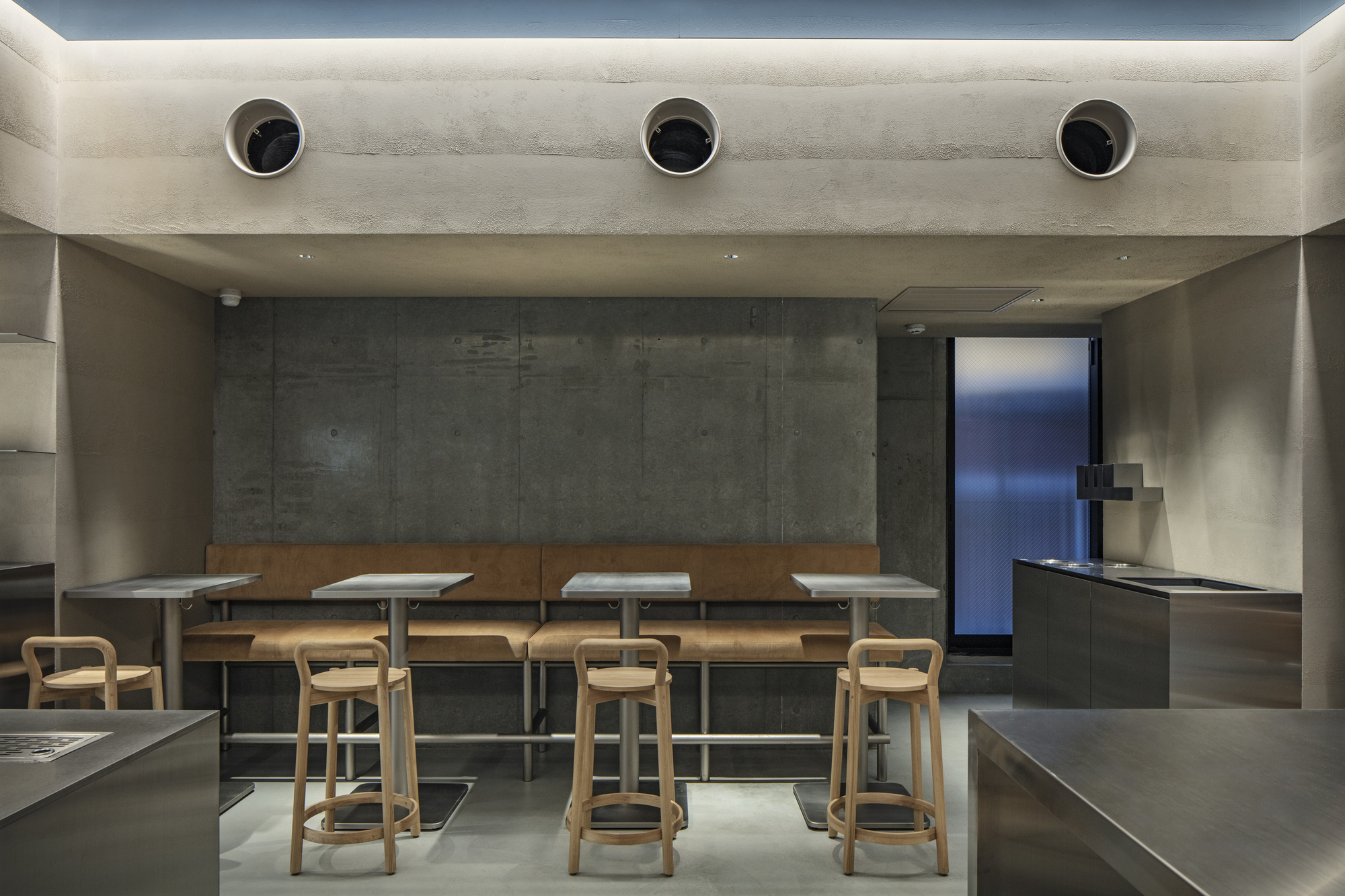 Seating area with tan leather banquette, steel-topped tables and wooden stools against a board-marked concrete wall under a softly lit ceiling