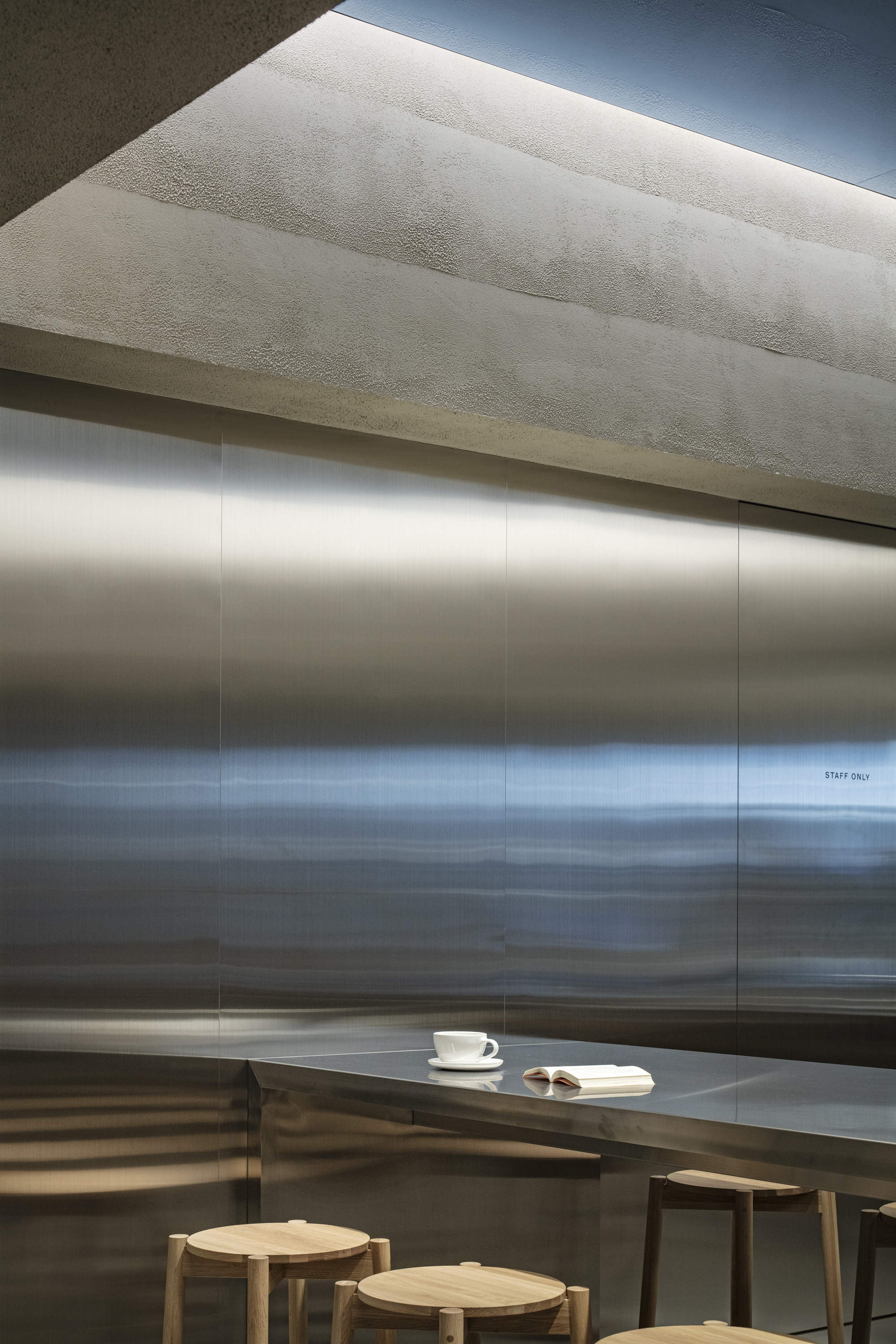 Brushed stainless steel wall above a counter holding a white cup and an open book, with wooden stools in the foreground