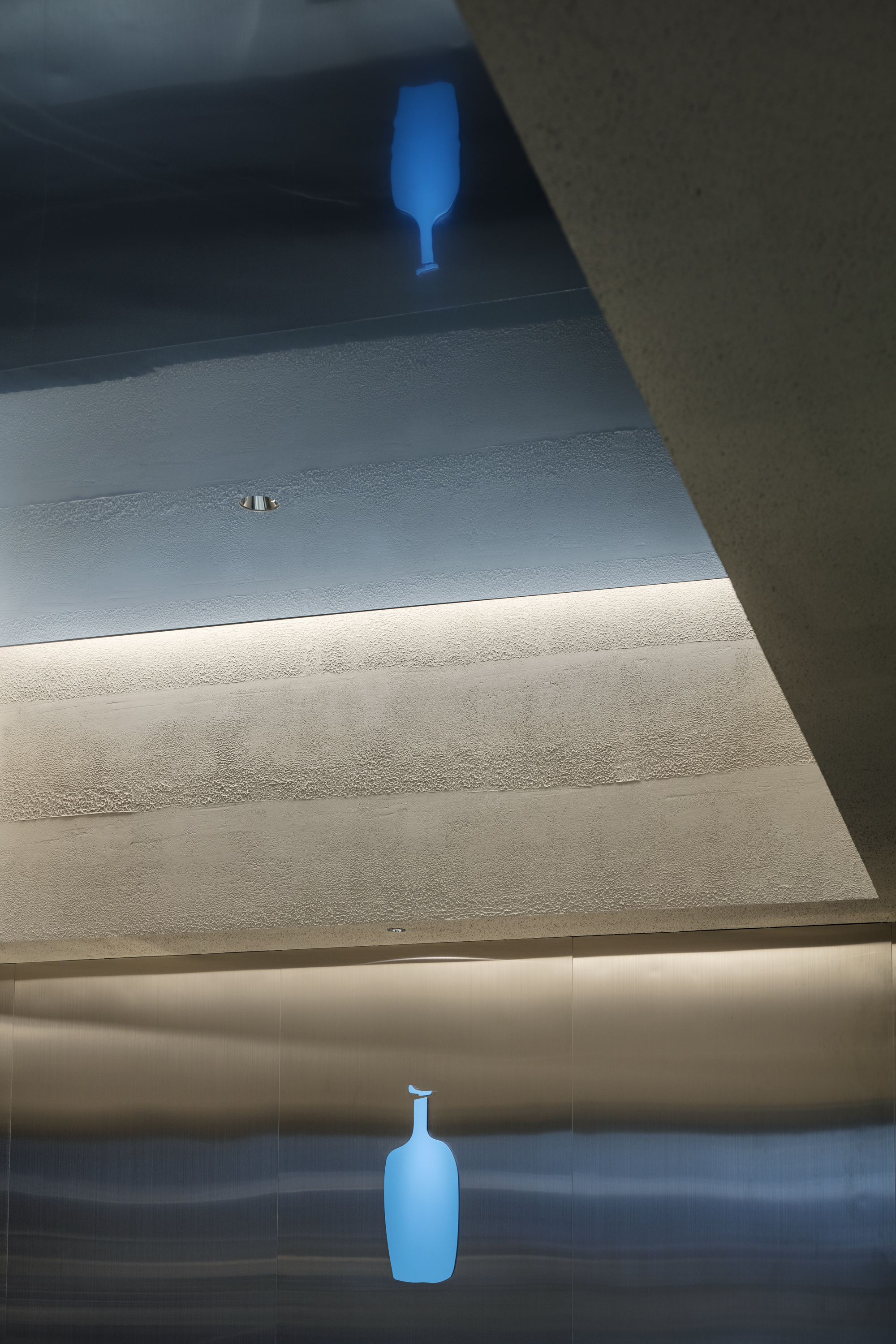 Blue bottle logo glowing on the brushed steel wall and reflected in the textured ceiling, lit by a cove light