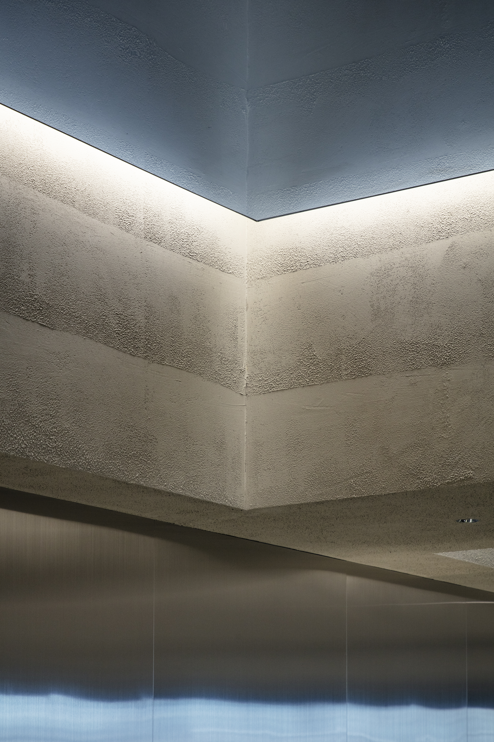 Corner of the layered rammed earth wall washed by a concealed cove light beneath the blue-grey ceiling, above a steel panel