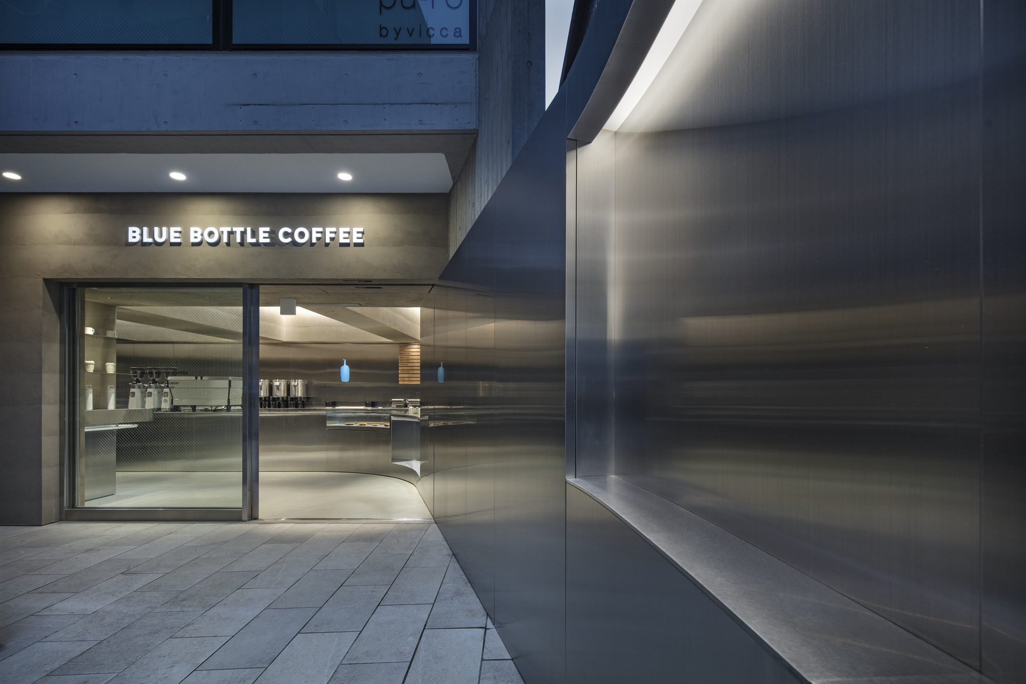 Glazed café entrance beside a curved stainless steel wall with a recessed light strip, seen at dusk