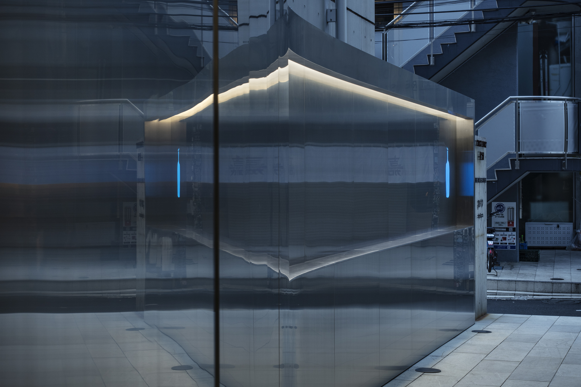 Corner of the brushed steel wall reflecting the street, with glowing blue bottle marks and a lit upper edge