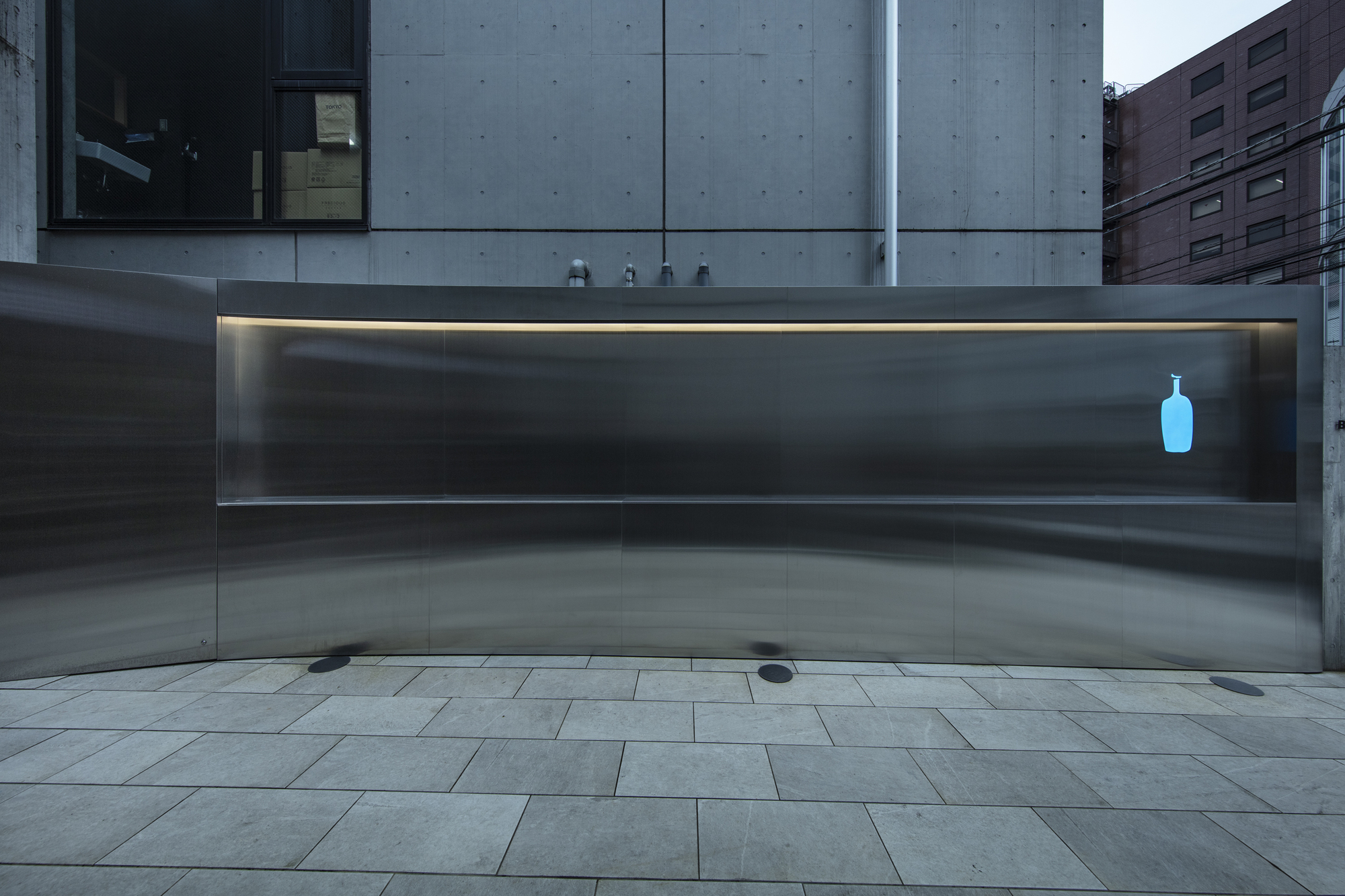 Frontal view of the curved stainless steel counter wall with a blue bottle logo and a concrete facade behind