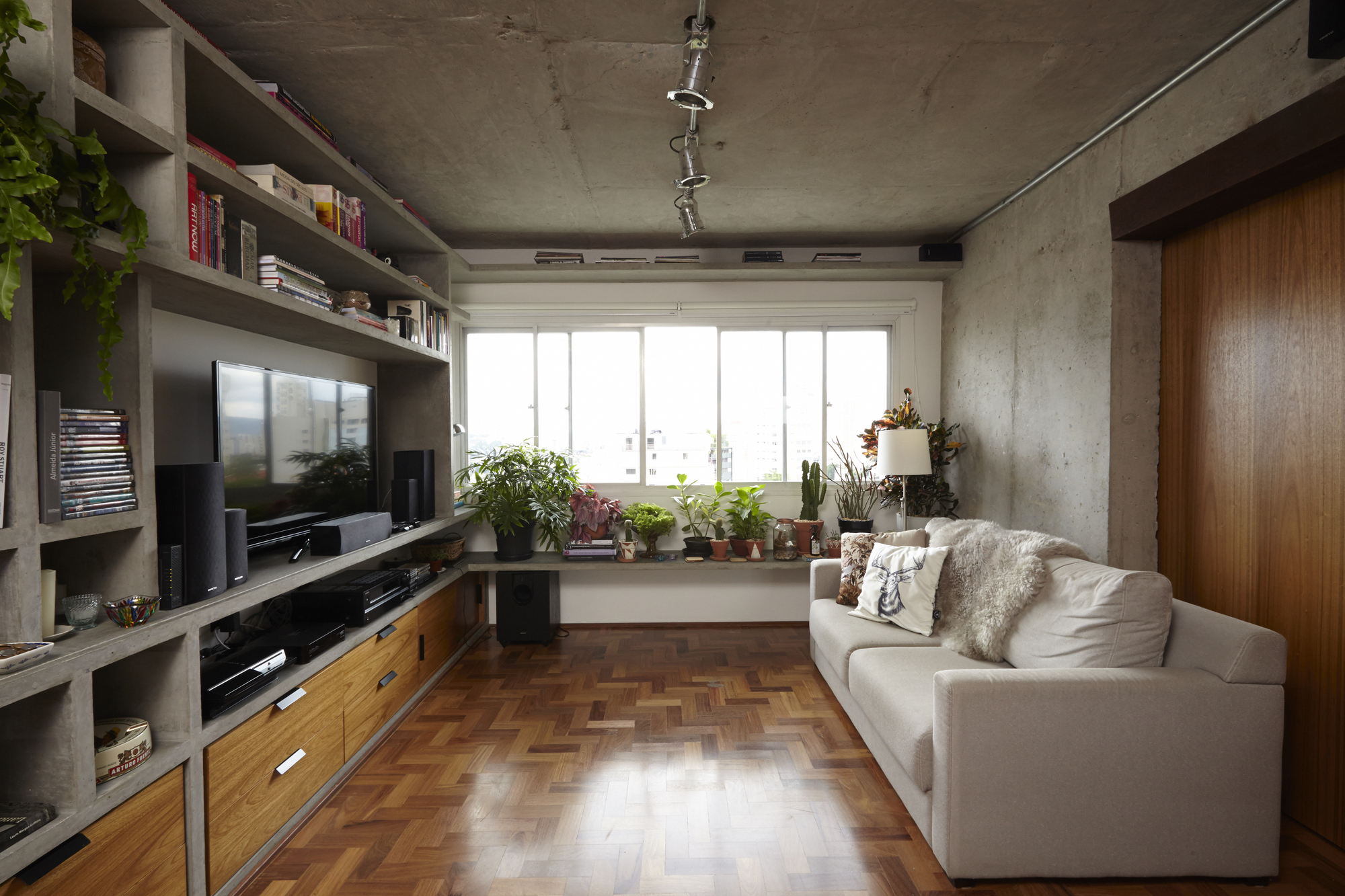 Living room with a concrete shelving unit, herringbone parquet floor and grey sofa facing a window lined with potted plants in daylight