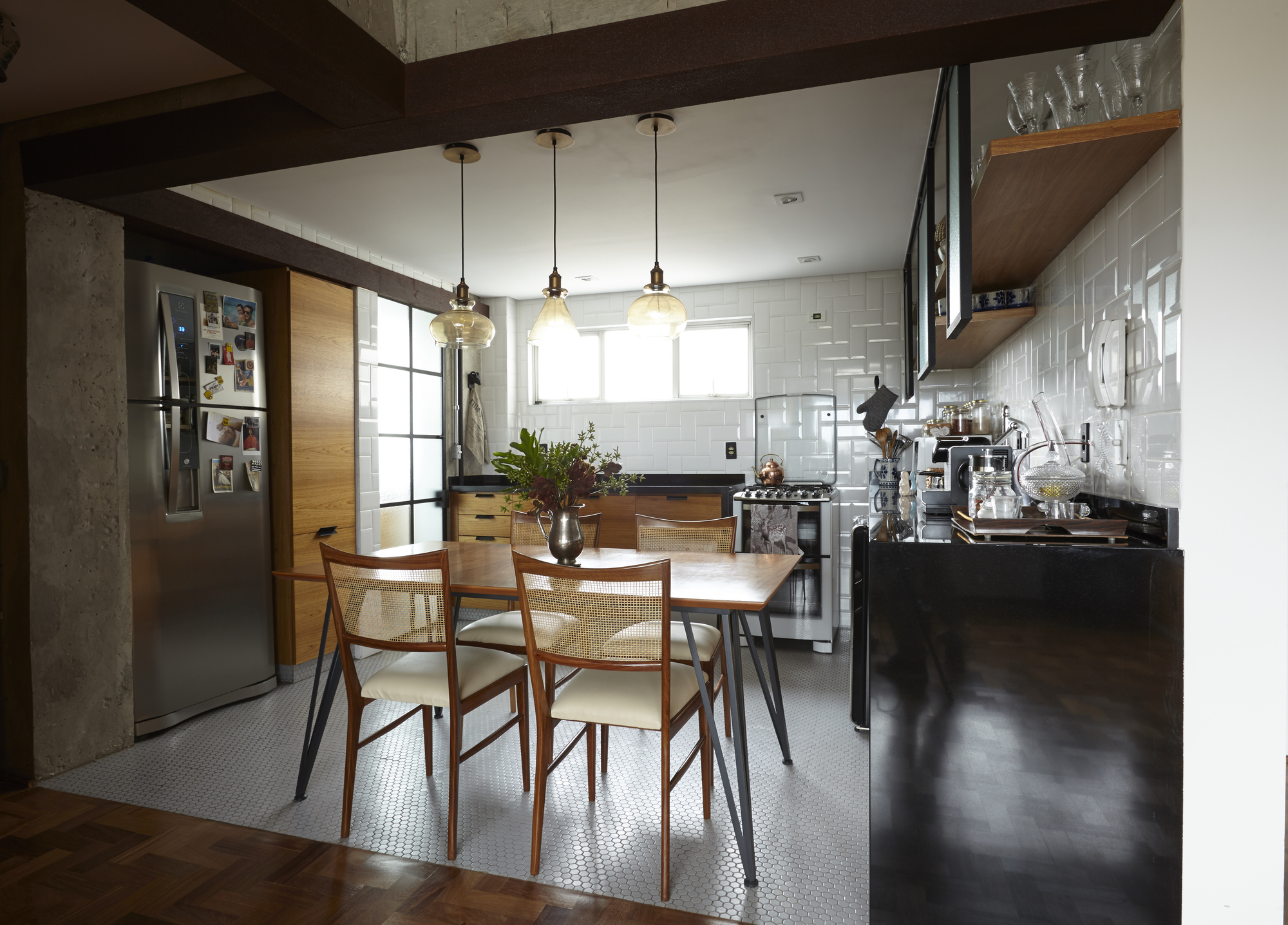 Kitchen seen from the parquet hallway, with a wooden dining table, cane-back chairs and glass pendant lamps over white tile and hexagonal flooring