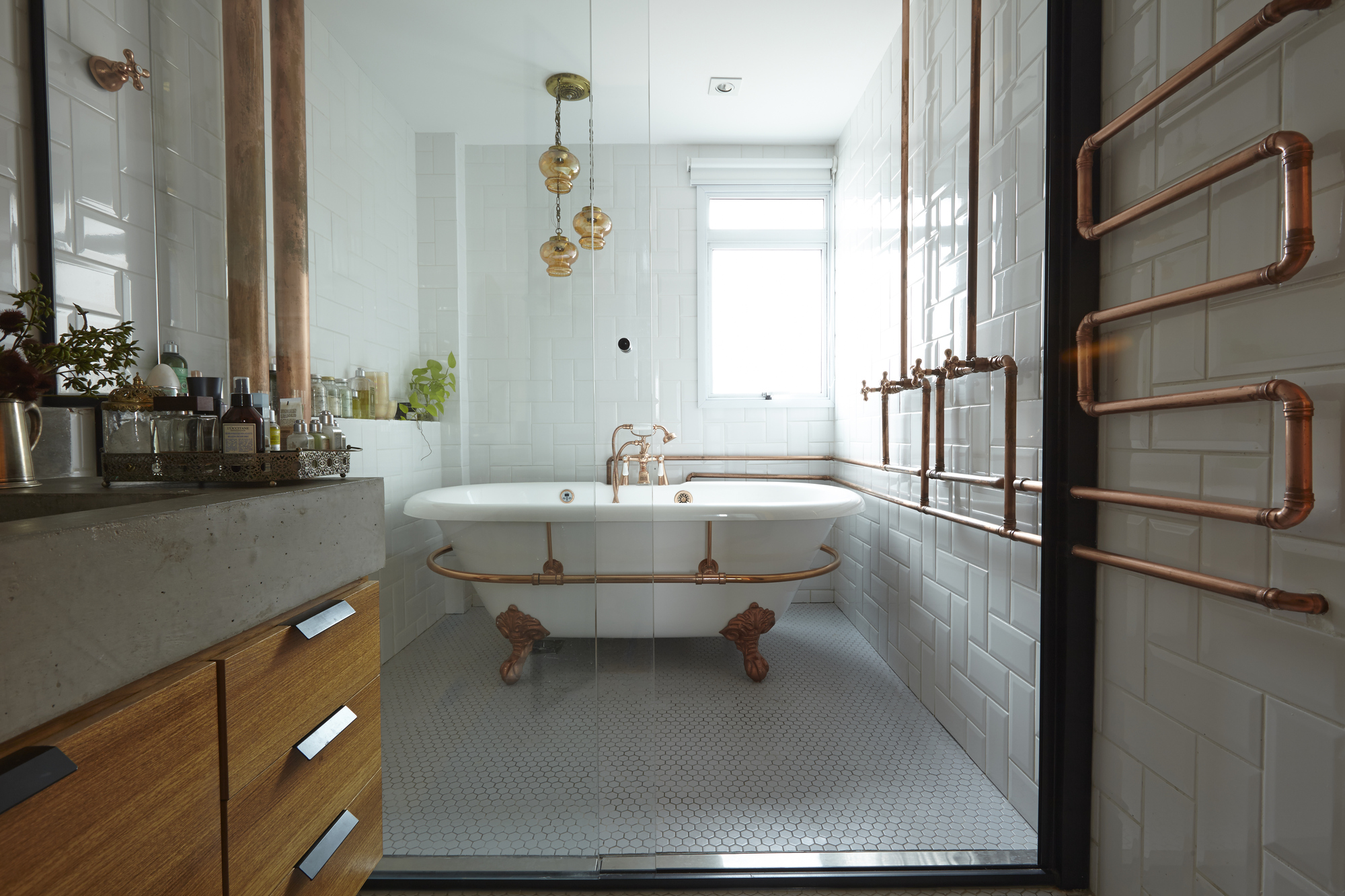 Bathroom with a freestanding clawfoot tub, exposed copper pipes and a towel warmer against white subway tile, lit by a bright window