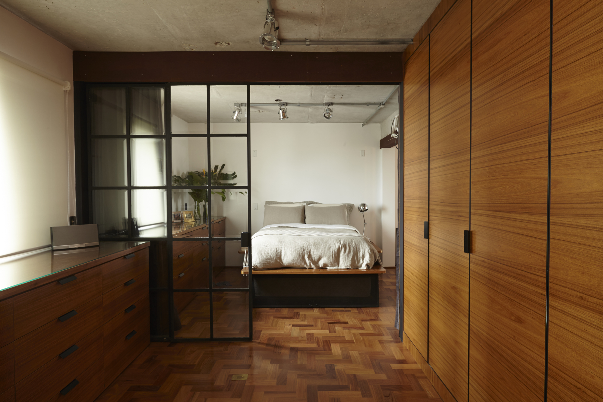 Bedroom seen through a black steel-framed glass partition, with a teak wardrobe wall and herringbone floor beneath a concrete ceiling