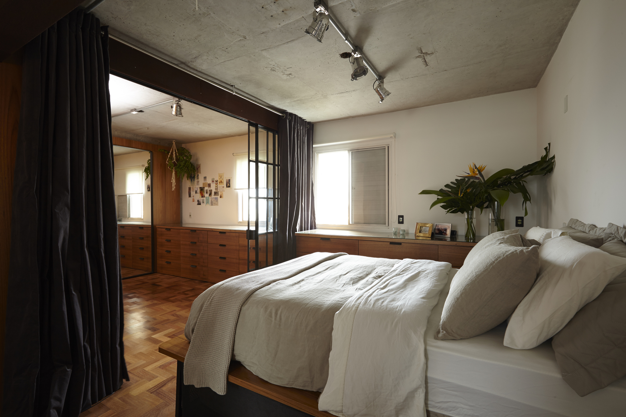 Bed with linen bedding beside a heavy dark curtain, with a bird-of-paradise plant on the dresser under track lights in daylight