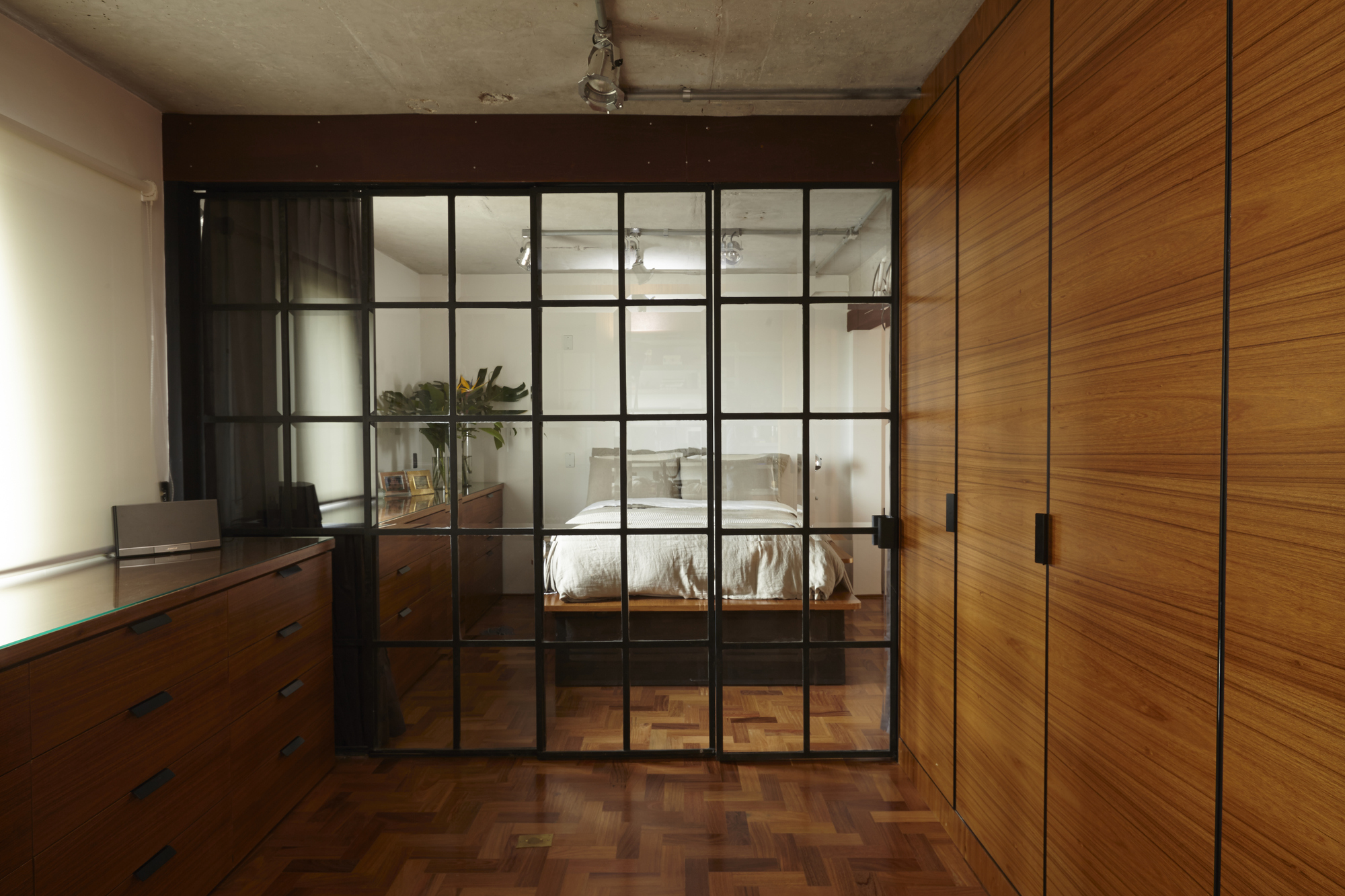 Black steel-framed glass partition between a teak-veneer wardrobe wall and a bedroom, with herringbone parquet floor and exposed concrete ceiling