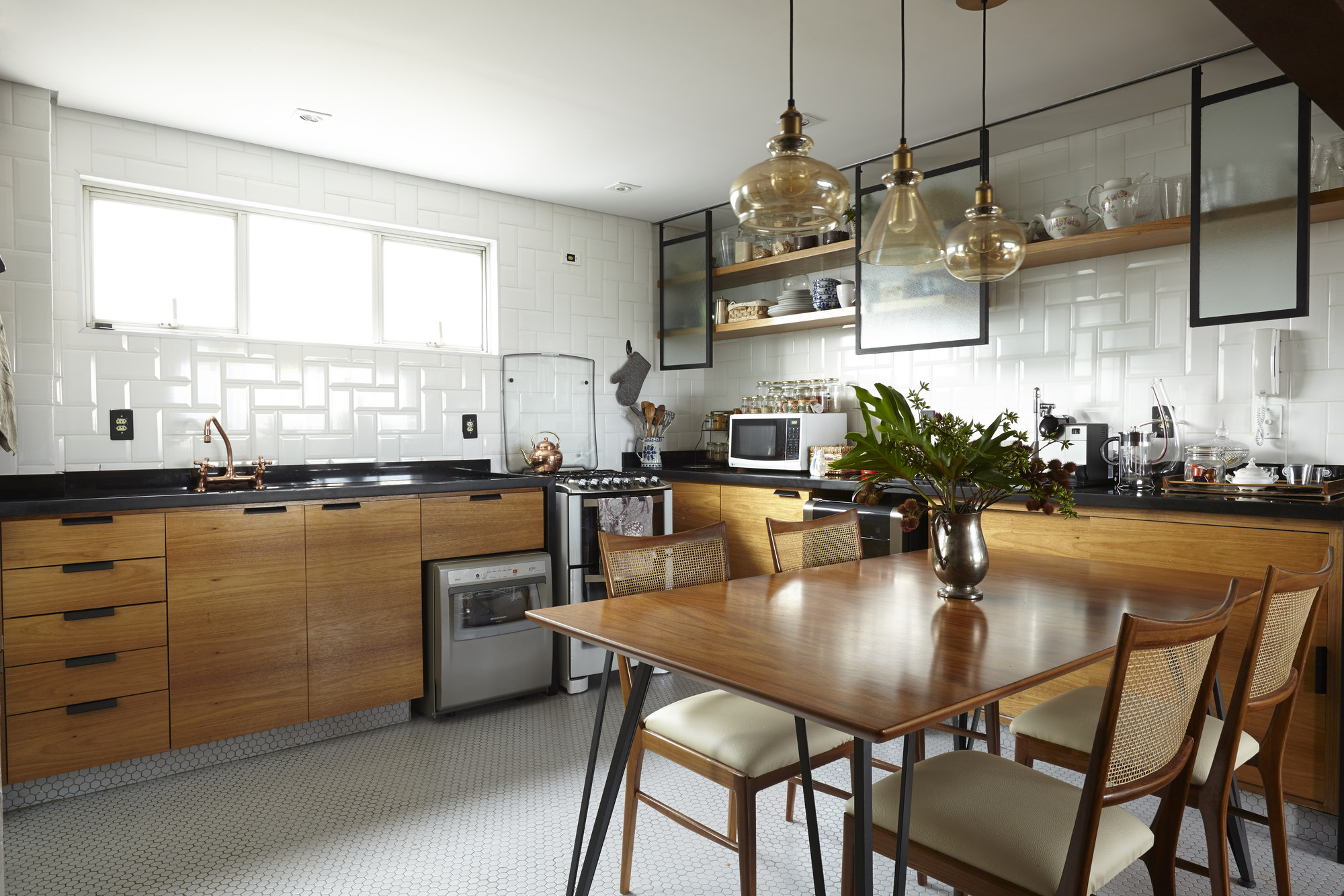 Kitchen with timber cabinets, black counters and white subway tile, lit by amber glass pendants over a cane-back dining table