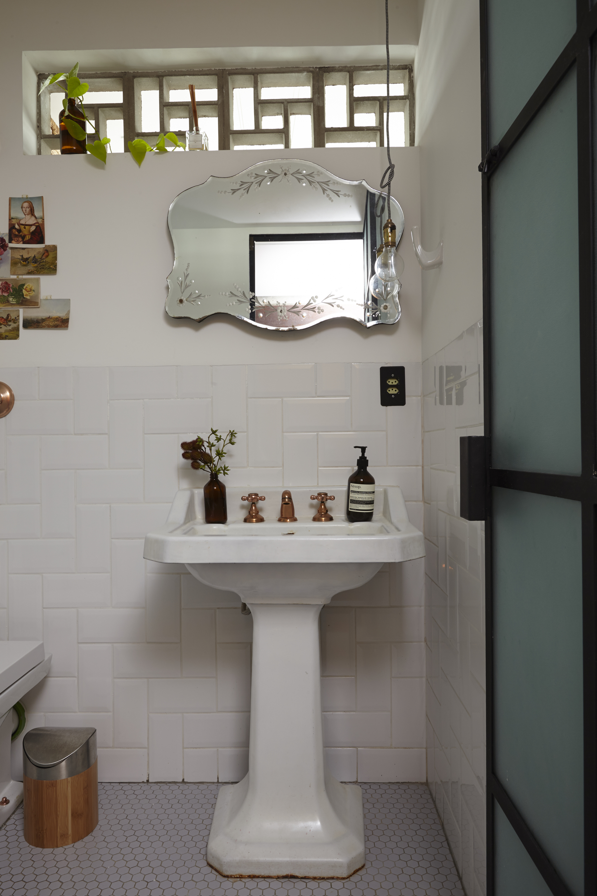 White pedestal basin with copper taps beneath a scalloped etched mirror and a high glass-block window with a trailing plant