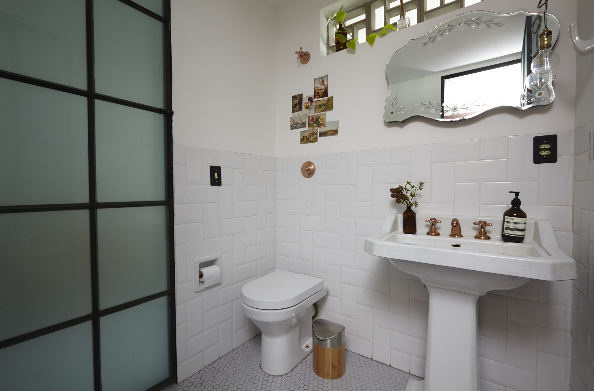 Bathroom corner with toilet, pedestal basin and frosted steel-framed door, daylight reflected in the etched mirror above the white tile