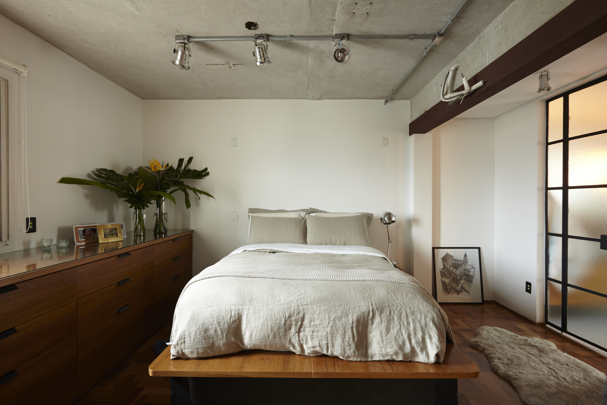Bed on a timber platform beside a long wooden dresser, with track lights on the raw concrete ceiling and a sheepskin rug
