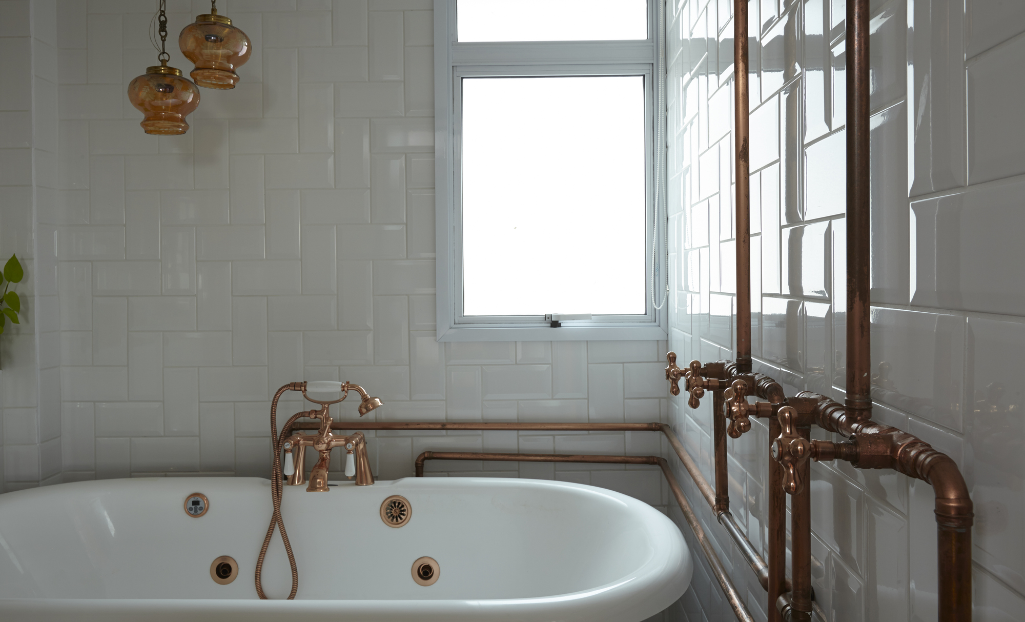Freestanding white tub beneath a bright window, with exposed copper pipes running along glossy white tiled walls and amber pendants