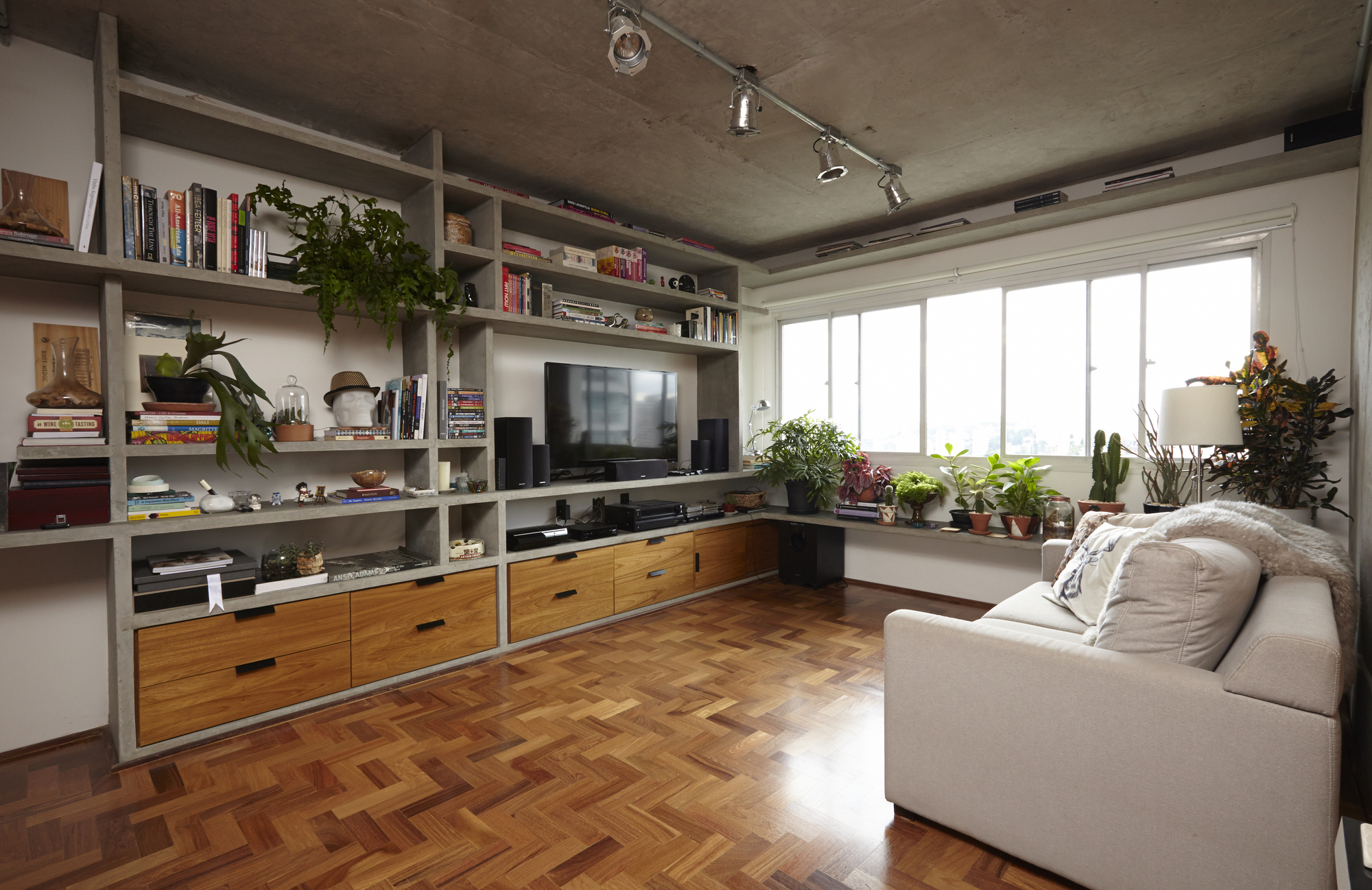 Living room with built-in shelving, wood drawer units and herringbone parquet floor, daylight streaming through a wide window with potted plants