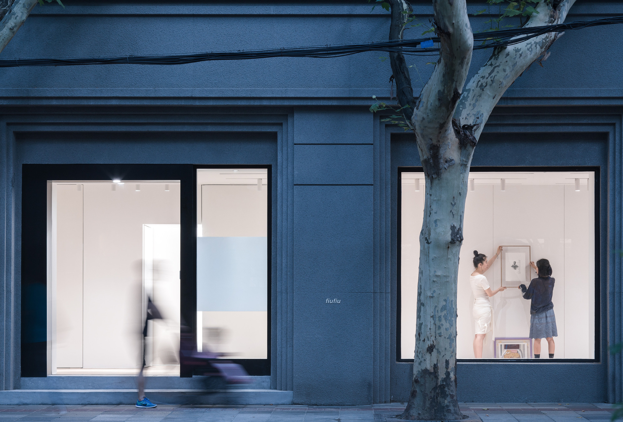 Blue-grey stone facade with two large shopfront windows, a plane tree trunk in front and two people hanging a framed picture inside at dusk