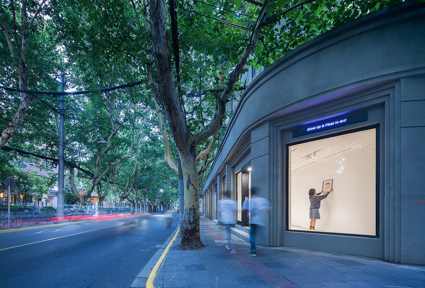 Curved corner of the grey facade with a lit window, blurred pedestrians and a row of plane trees along the street at dusk
