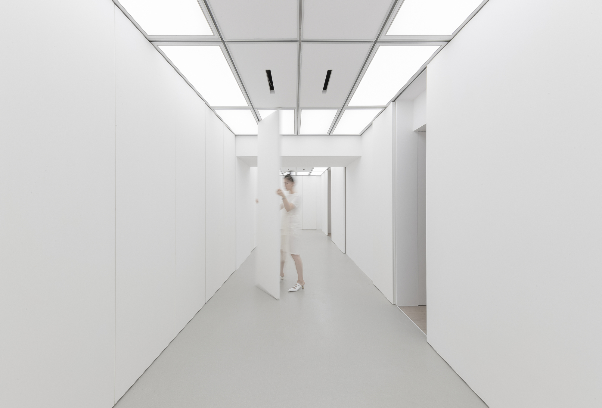 White corridor with luminous ceiling panels and a pivoting wall panel, a blurred figure in white standing beside it