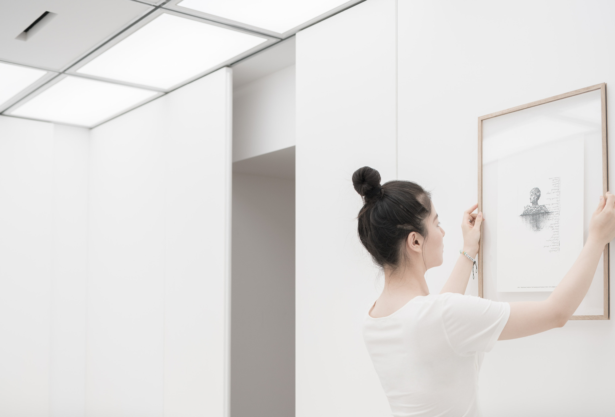 Woman with a hair bun hanging a framed drawing on a white wall beneath glowing ceiling panels