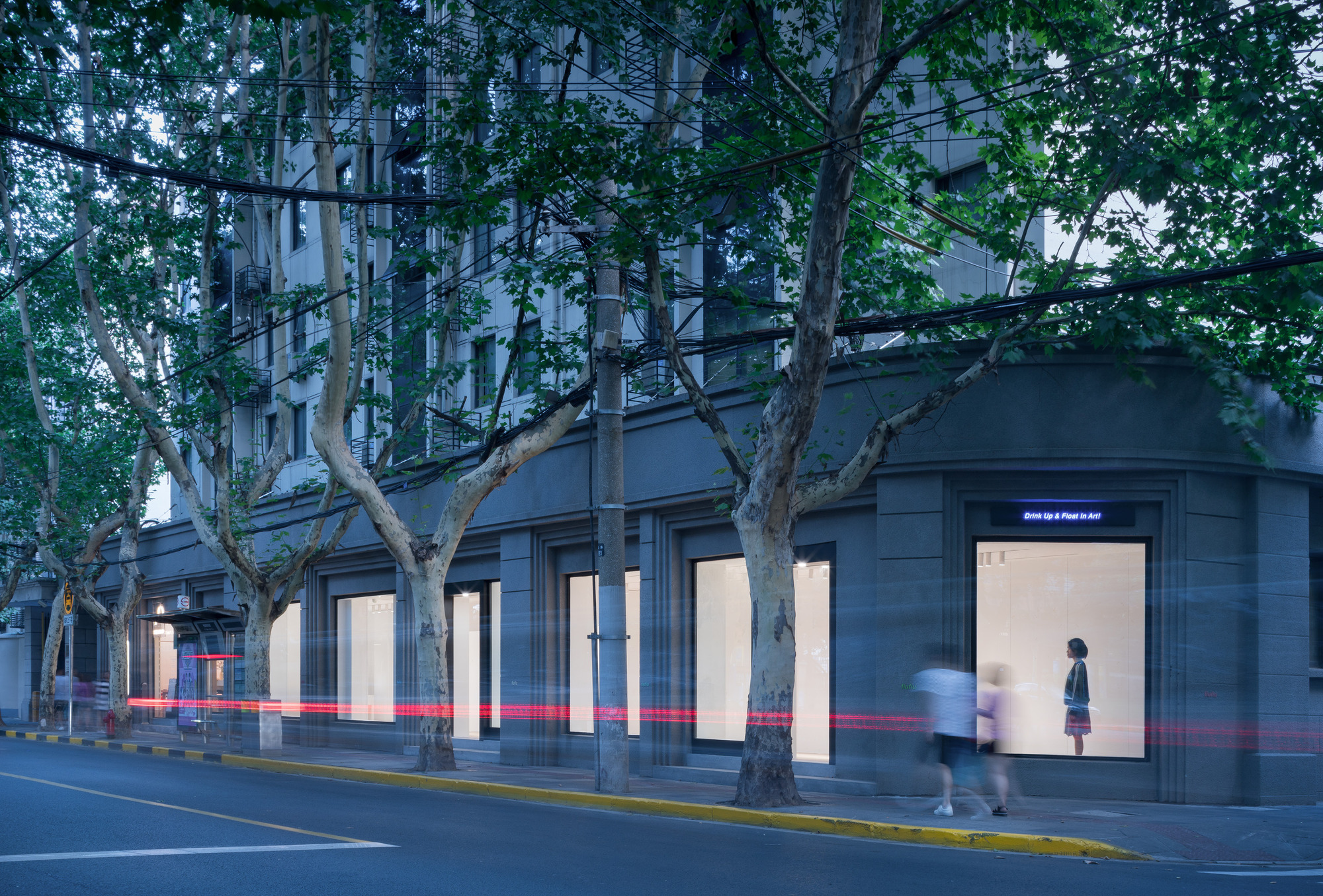 Corner storefront with tall lit display windows behind plane trees at dusk, a blurred pedestrian and red light trails crossing the pavement