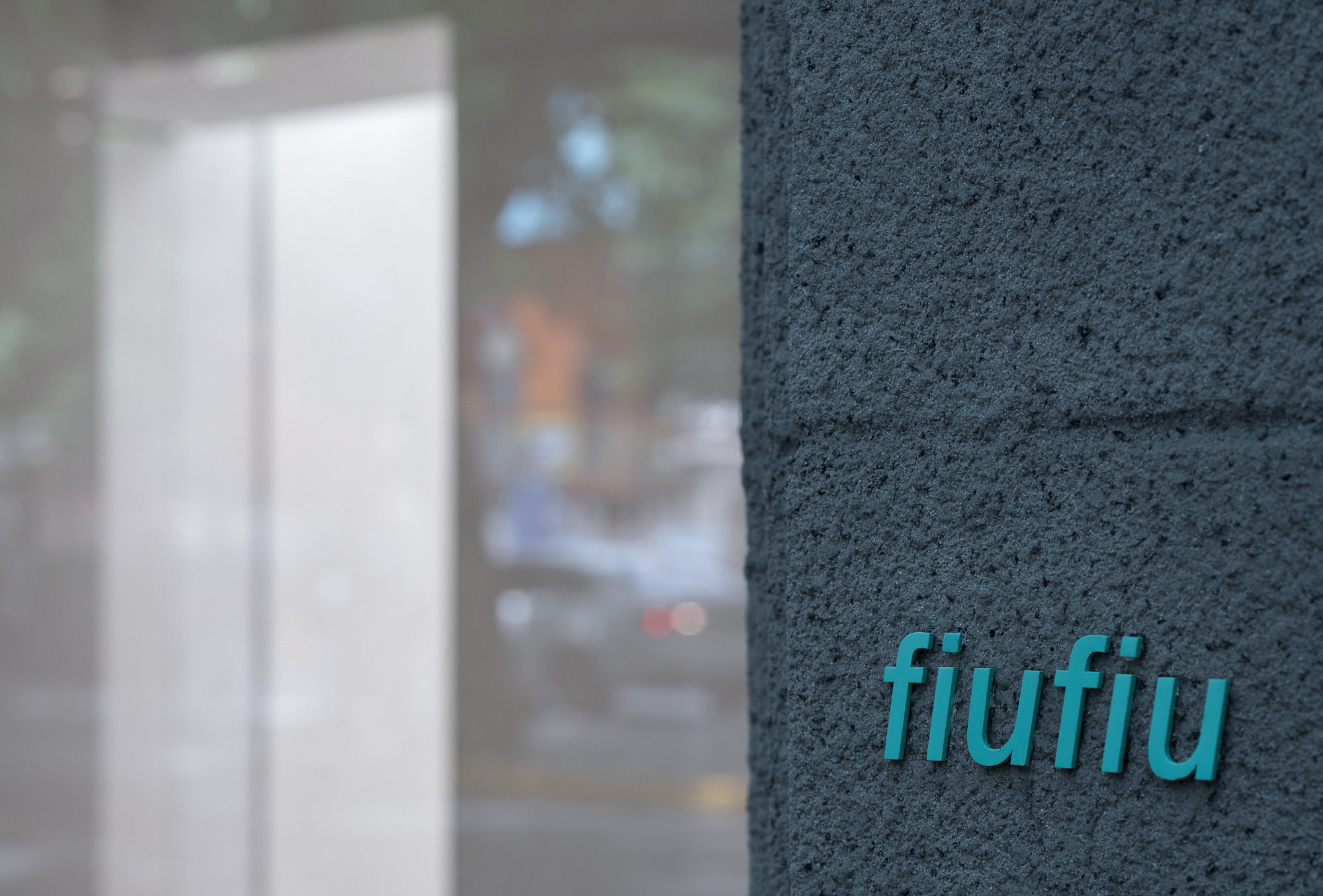 Close-up of teal lettering on a rough dark grey stone pier, with a blurred glass window and street behind