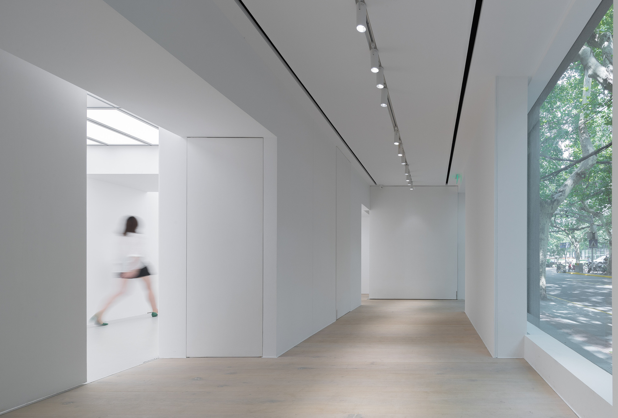 White gallery corridor with track lighting and oak floor, a blurred visitor stepping through a doorway beside a tall street window