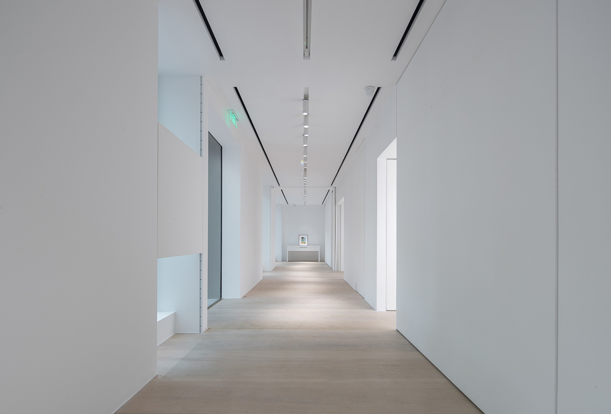 Long white corridor with recessed ceiling slots and pale timber floor, leading to a small framed artwork on a plinth