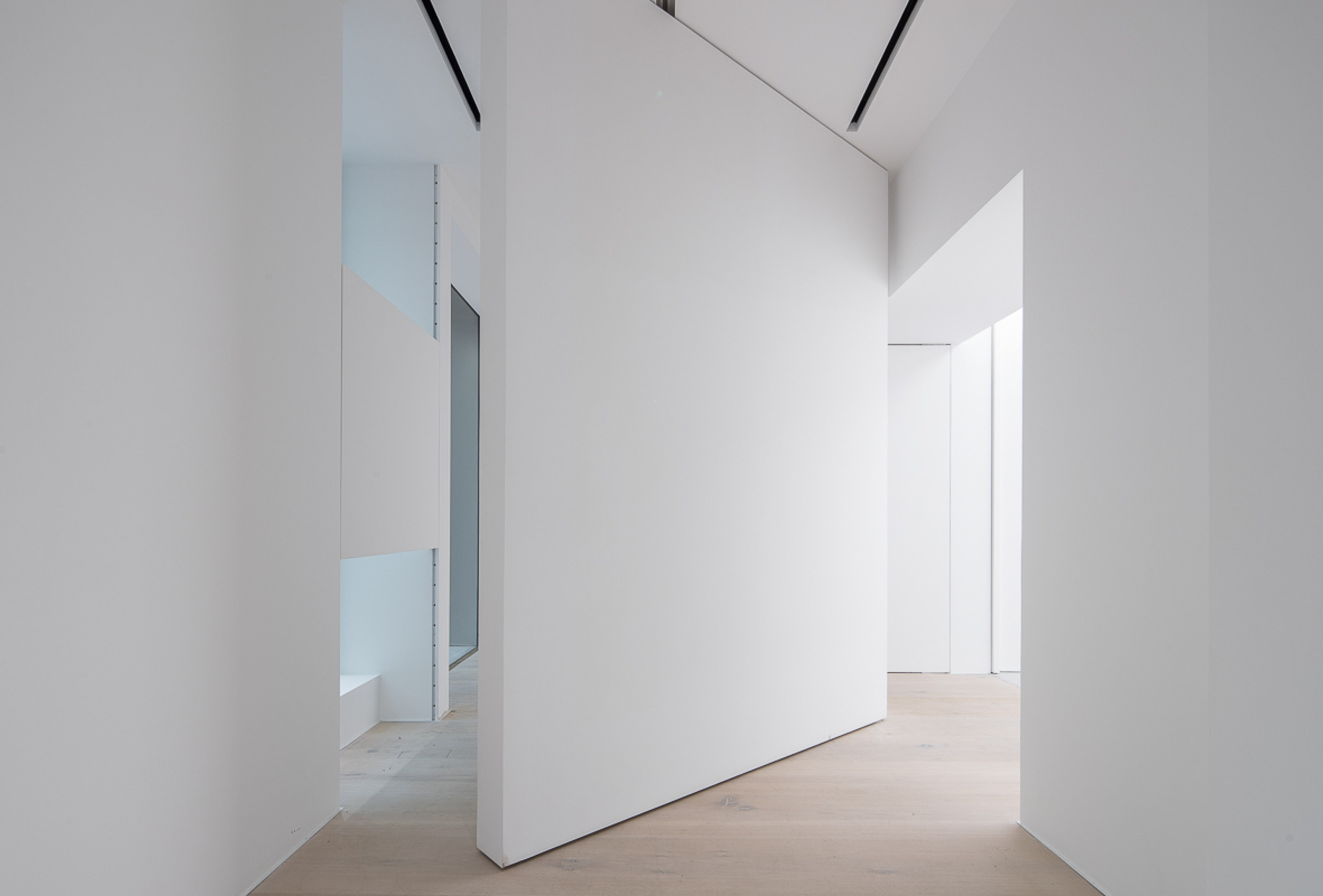 Freestanding white pivot wall angled between openings, with pale oak floor and slim ceiling light slots overhead