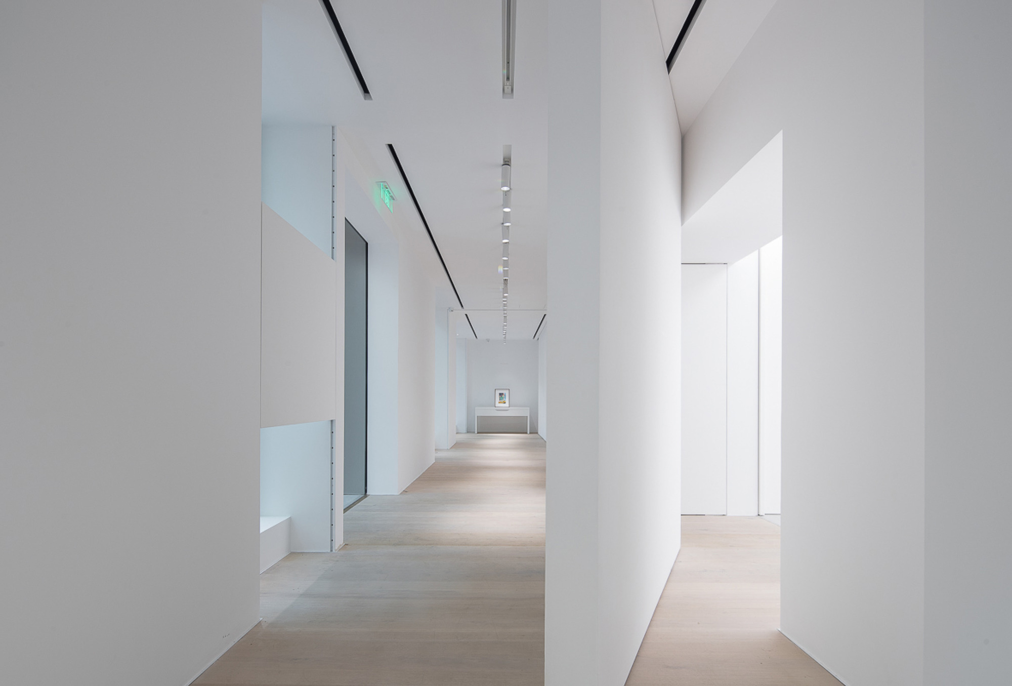 Gallery corridor with staggered white partition walls, pale timber floor and a small framed artwork on a plinth at the far end