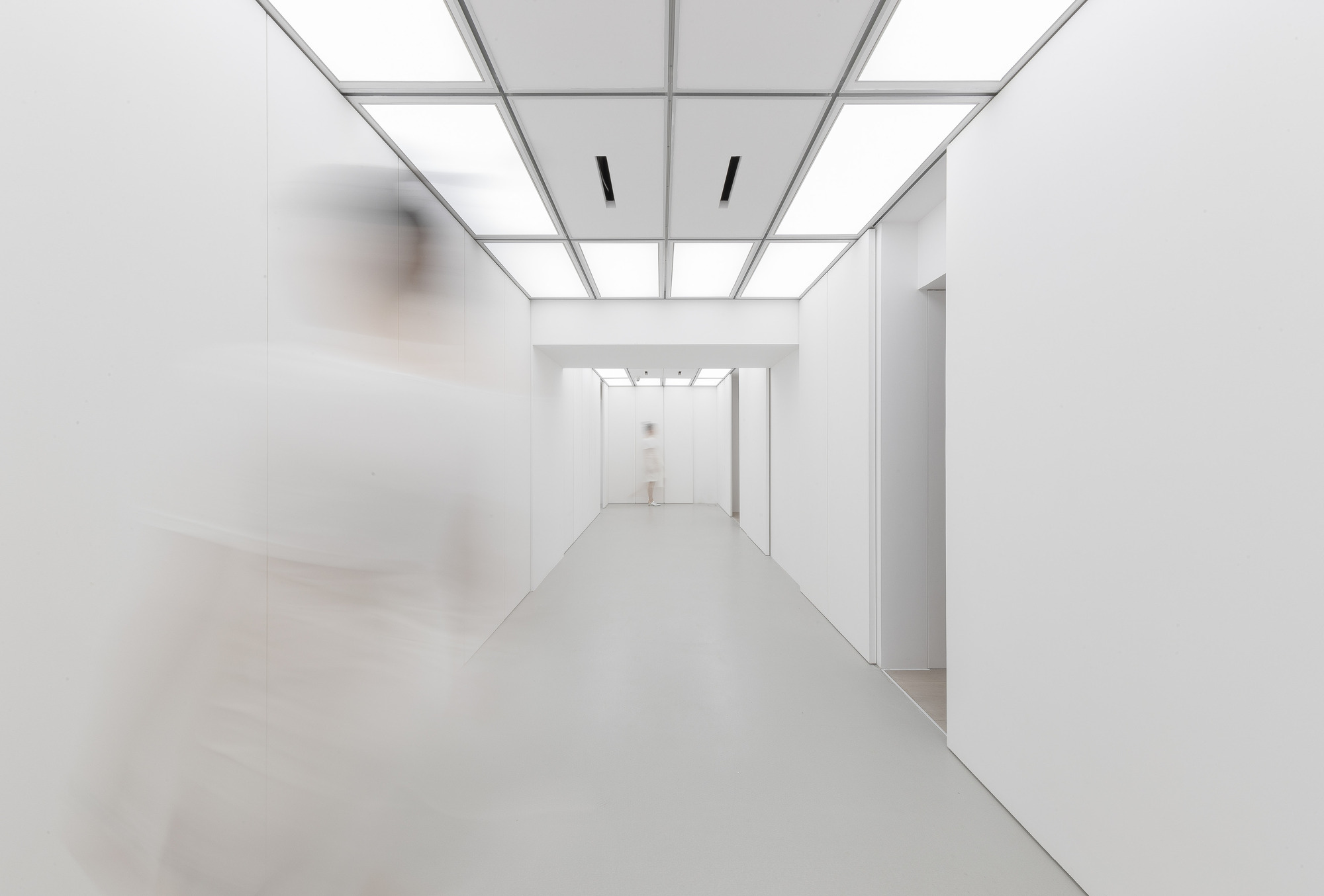Narrow white corridor with a luminous gridded ceiling and a blurred figure walking past in the left foreground