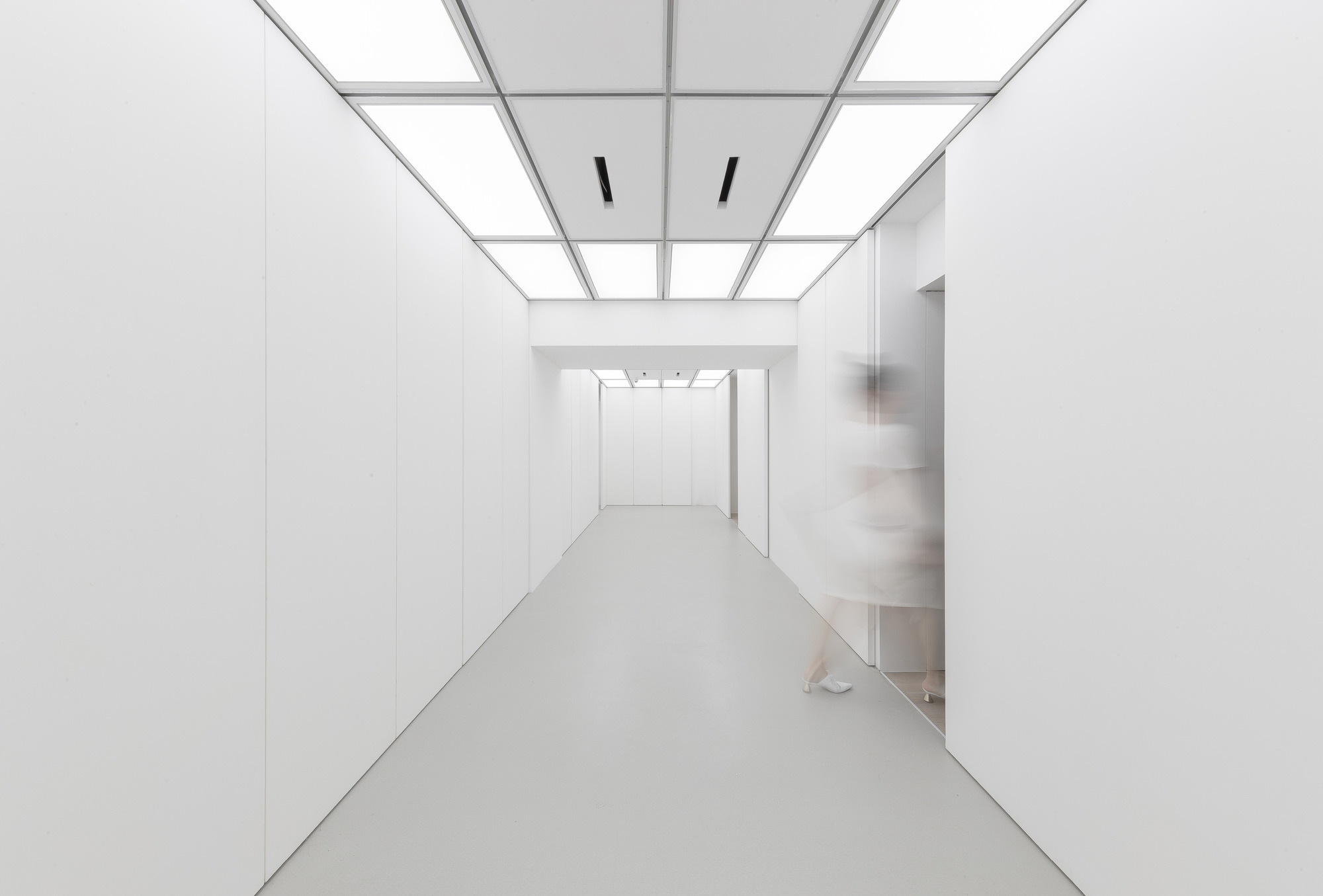 Same white corridor with illuminated ceiling panels and a blurred figure stepping through a doorway on the right