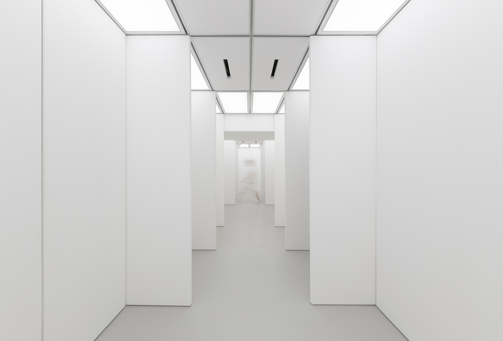 Row of freestanding white wall panels flanking a corridor, with a faint blurred figure walking in the distance