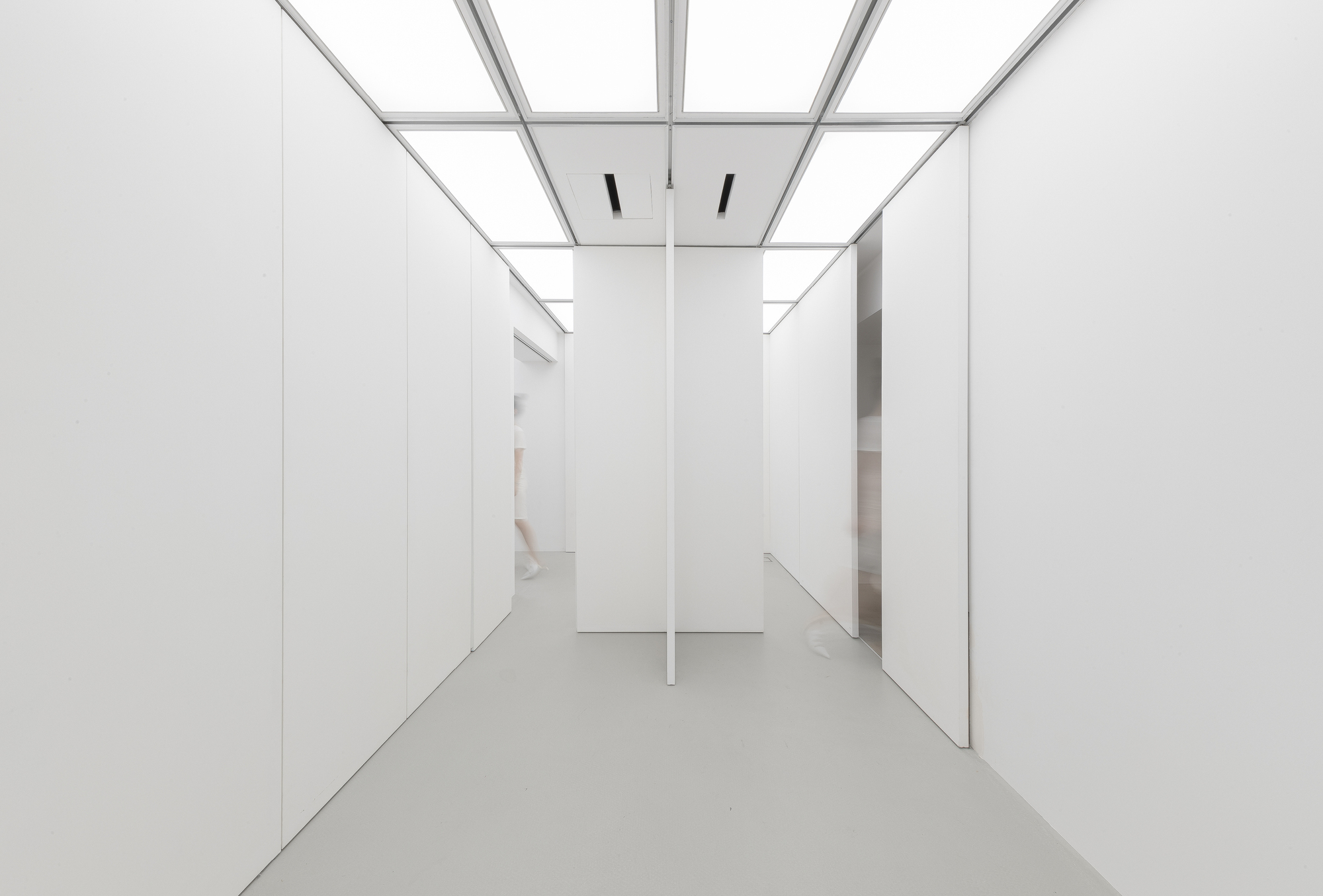 Corridor junction with a central white column, a thin vertical divider and a blurred figure passing behind the left wall