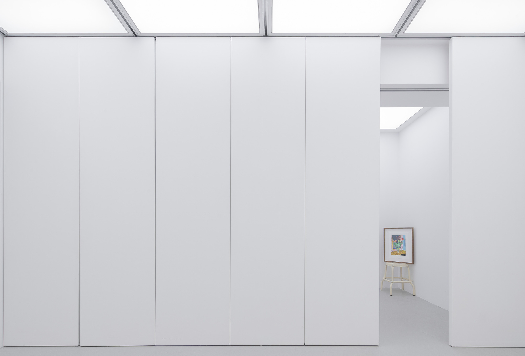 Row of tall white panel walls under luminous ceiling grids, with an opening framing a small painting on a stool