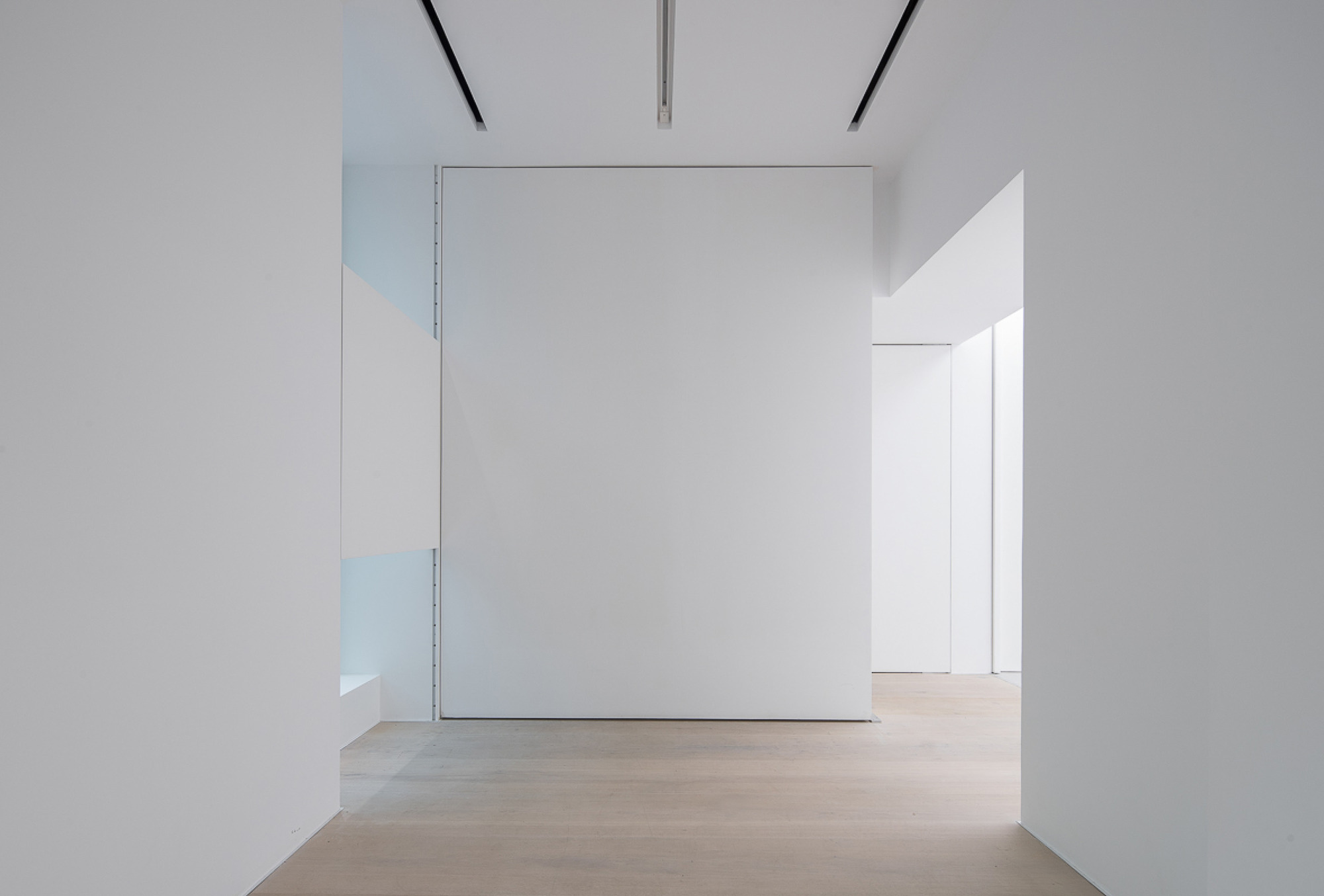 Gallery room with a freestanding white partition wall, pale oak floor and slim black linear ceiling slots in soft daylight