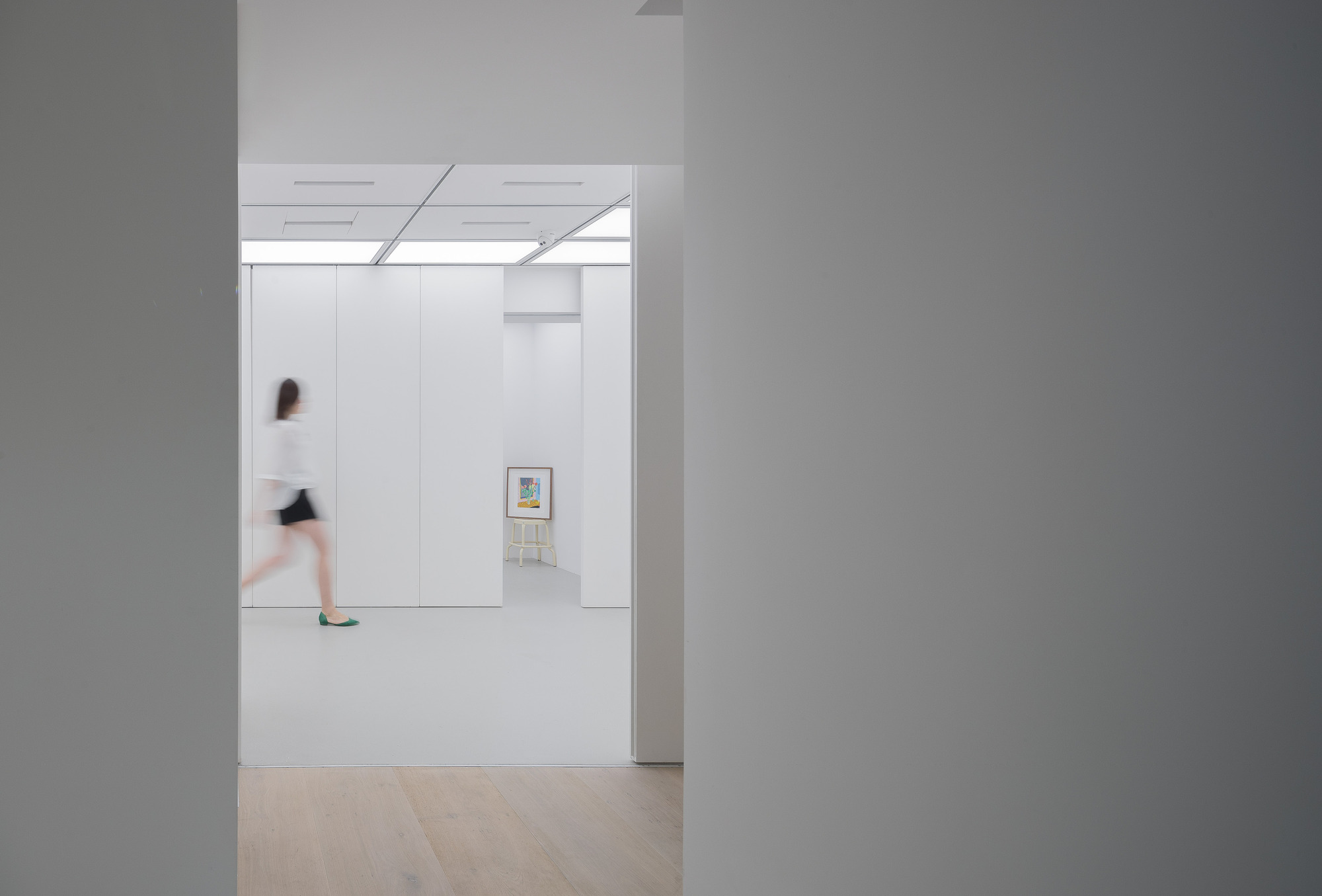 Blurred visitor walking past a doorway toward a framed picture on a stool, white panels and glowing ceiling beyond