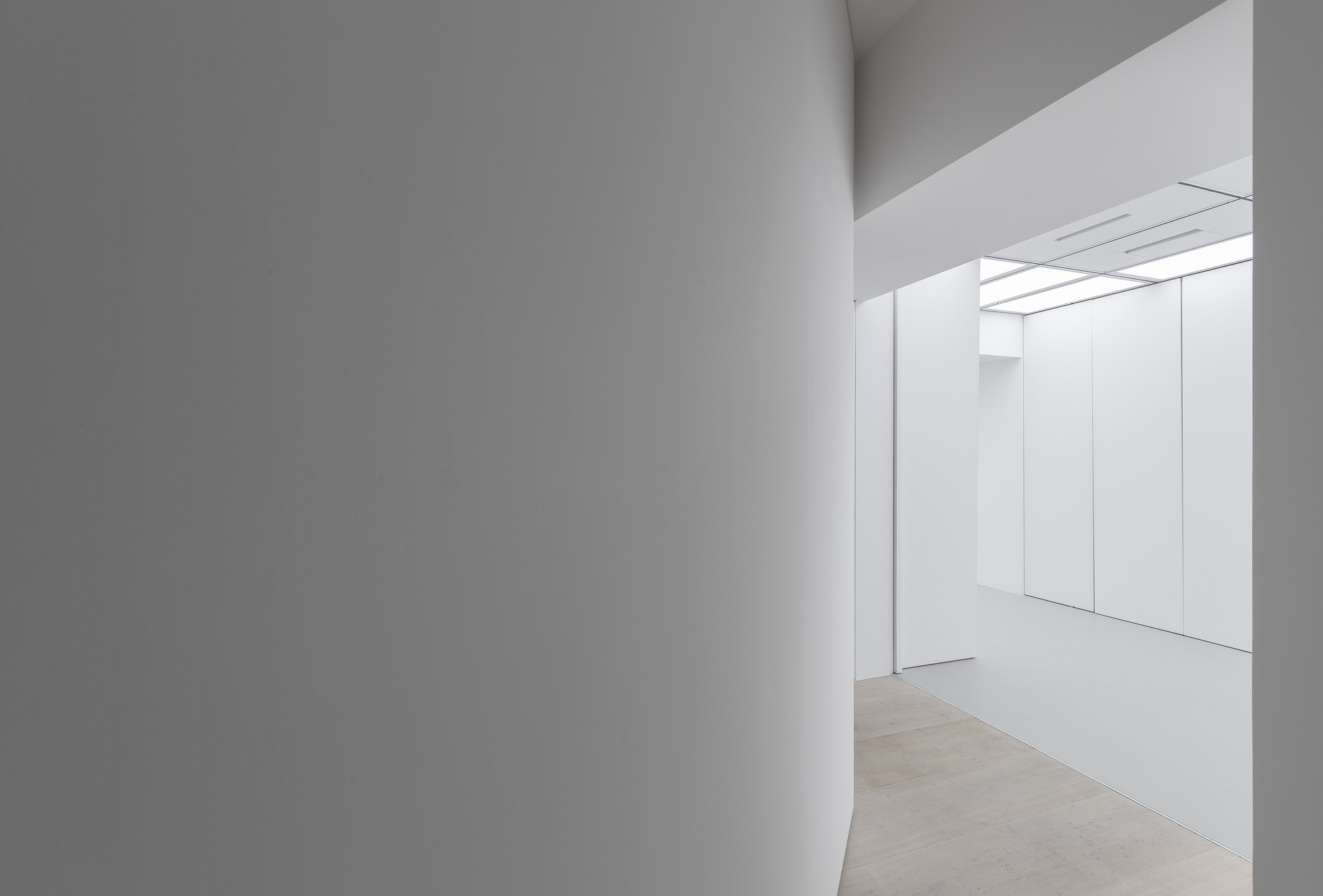 Curved white wall in the foreground leading to a brighter gallery with panelled walls and a luminous ceiling